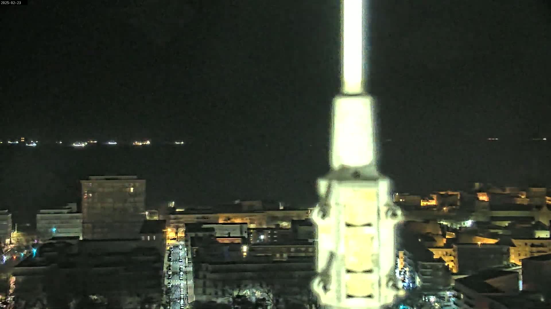 A nighttime, high-angle view shows a city's lights and a body of water with distant lights, partially obscured by a tall, light-colored structure in the foreground.