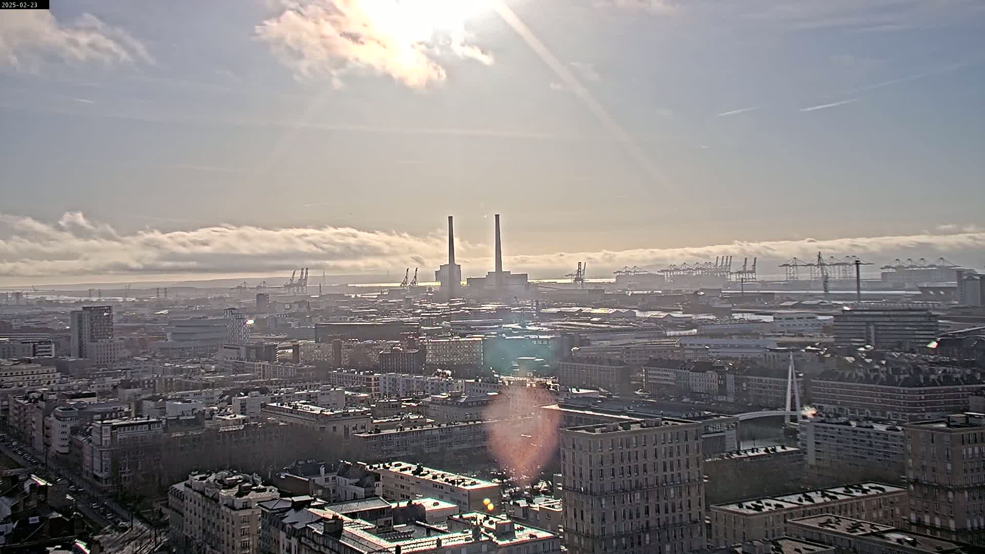 A hazy, sunny day reveals a sprawling cityscape with a distant port filled with cranes and two prominent industrial smokestacks.