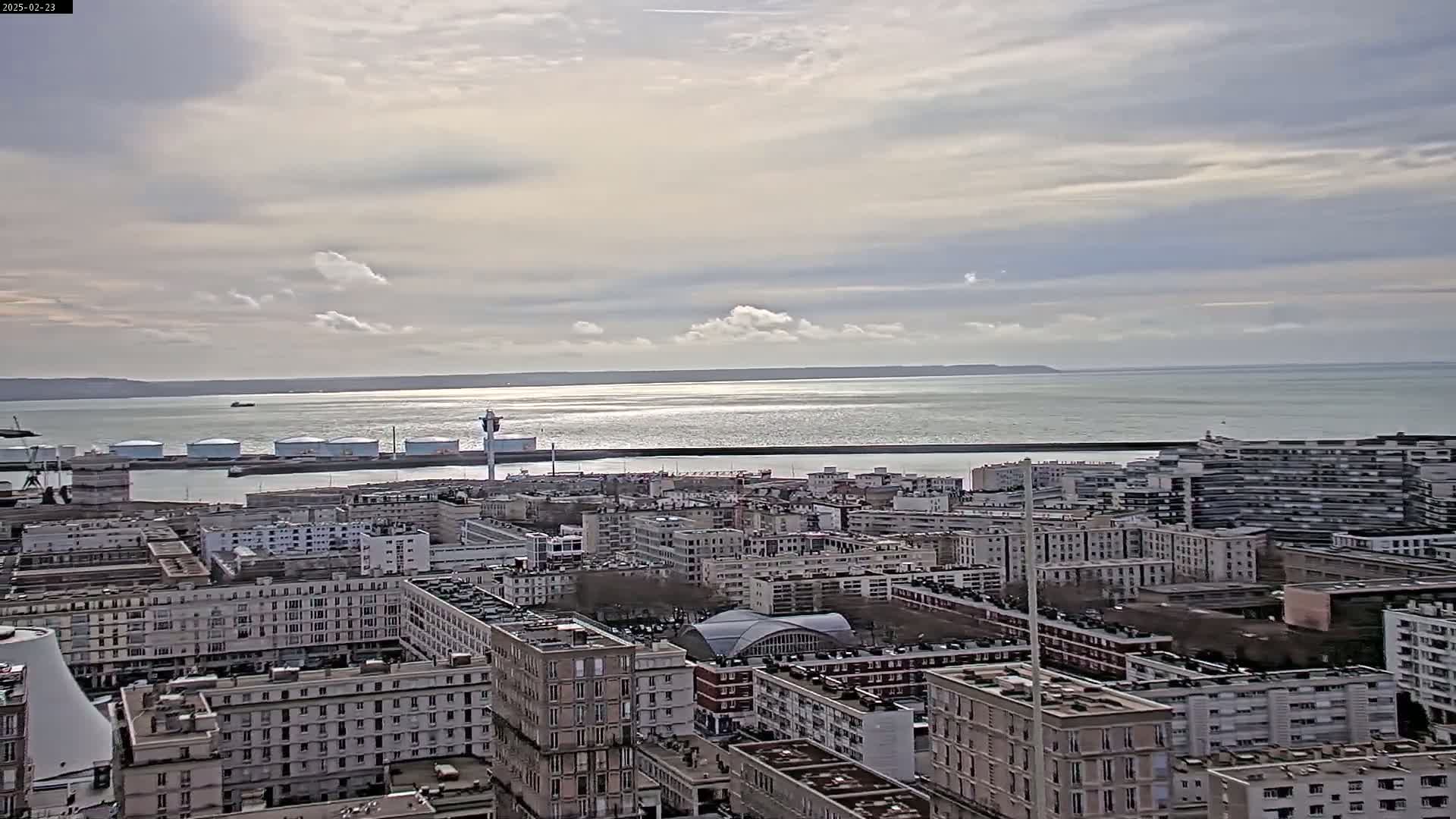 A sprawling cityscape overlooks a calm body of water under a partly cloudy sky.