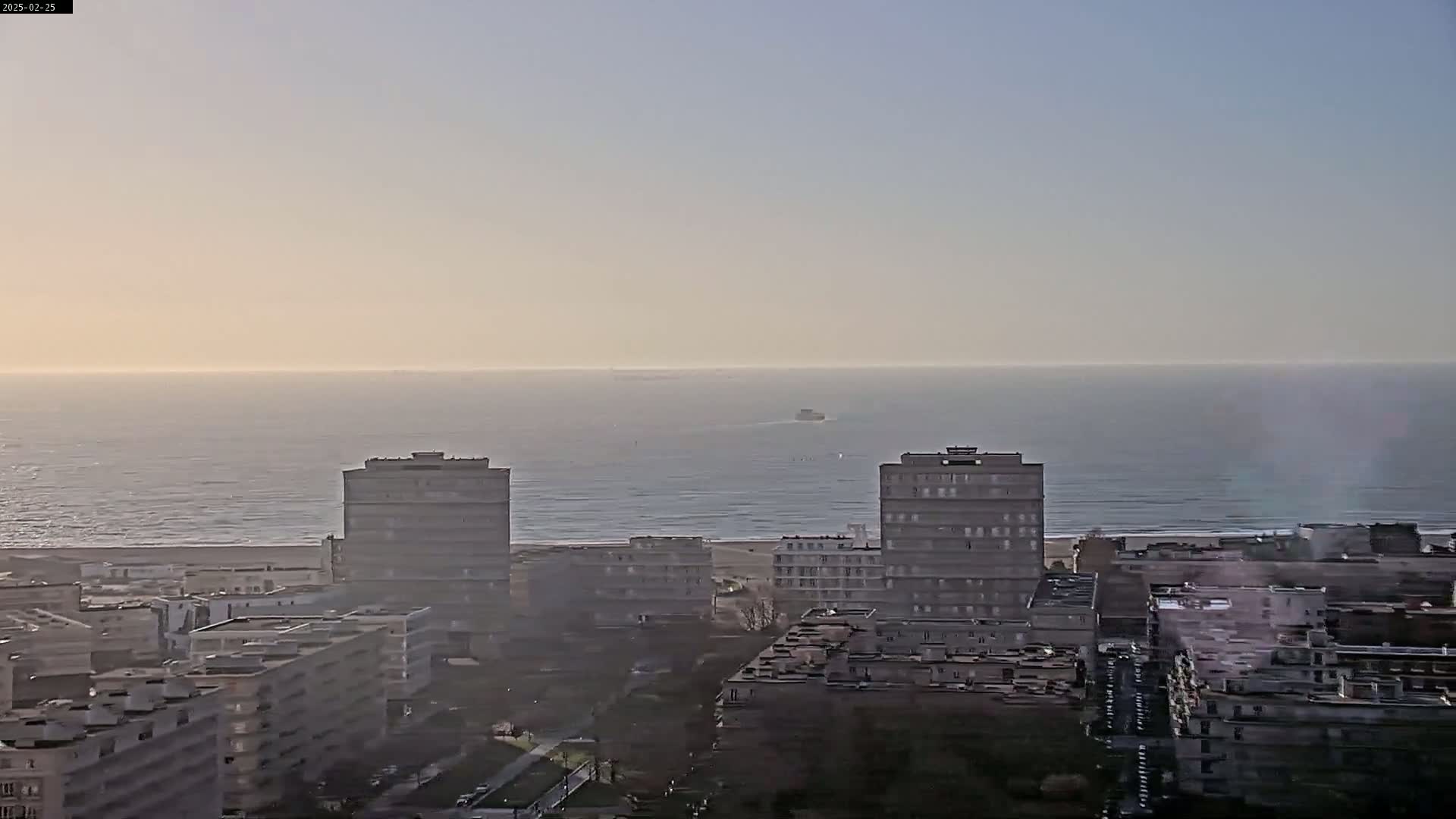A hazy sunrise illuminates a cityscape of multi-story buildings overlooking a calm ocean with a few distant ships.