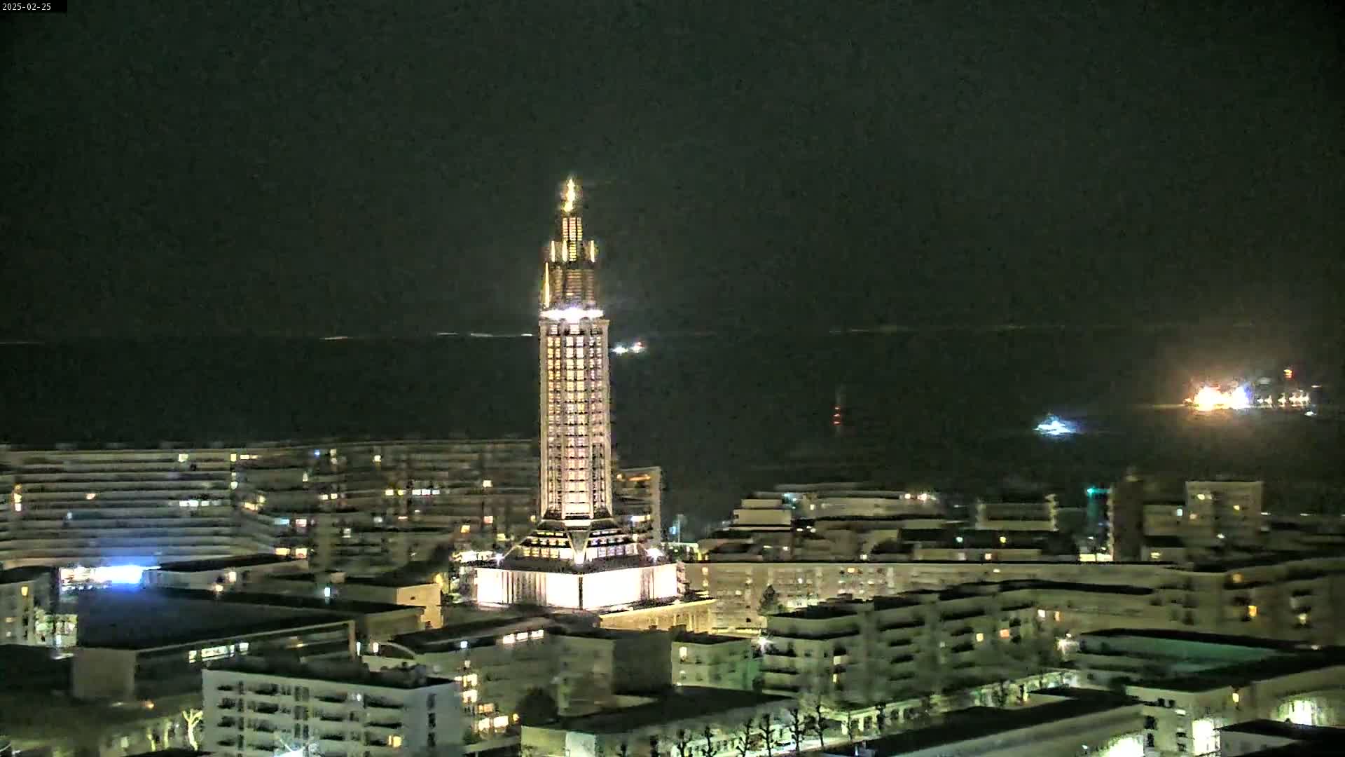 A tall, illuminated tower stands in the center of a city at night, overlooking a dark body of water.