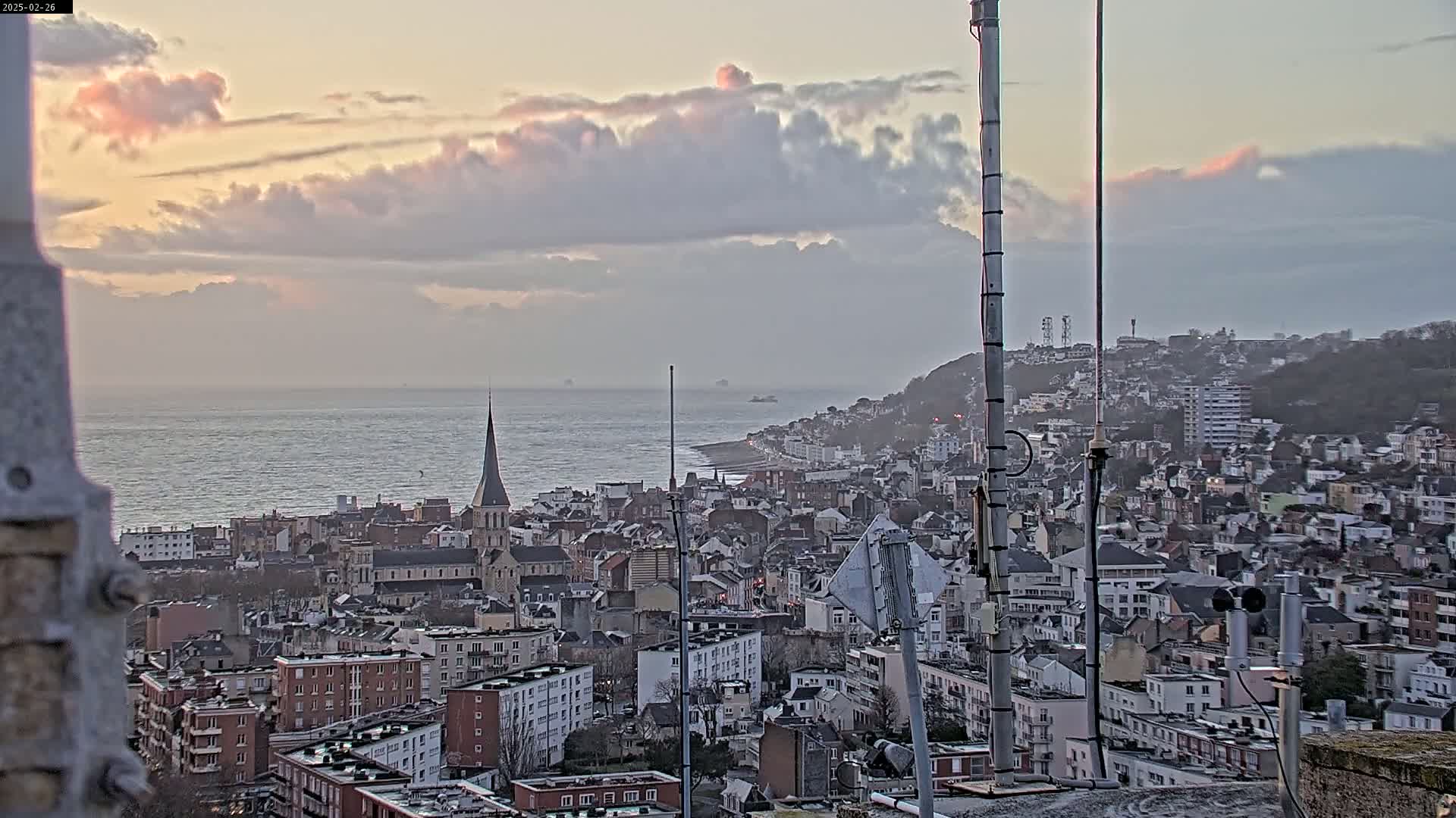 A coastal city with a church spire is visible under a cloudy, pink-tinged sky, with a calm sea in the background.