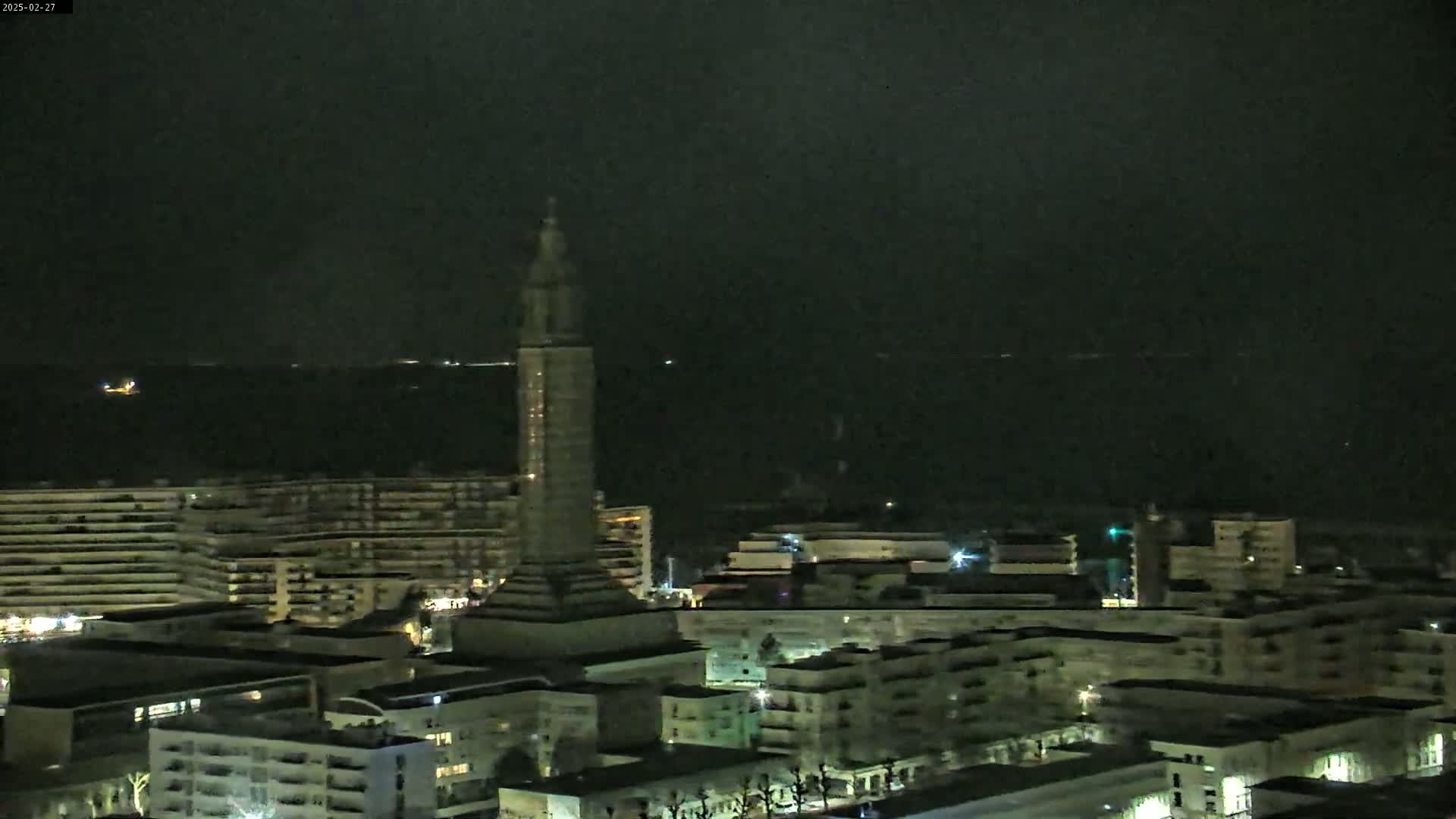 A nighttime view of a city with a tall, slender tower, faintly lit buildings, and a dark body of water in the distance under a mostly clear sky.
