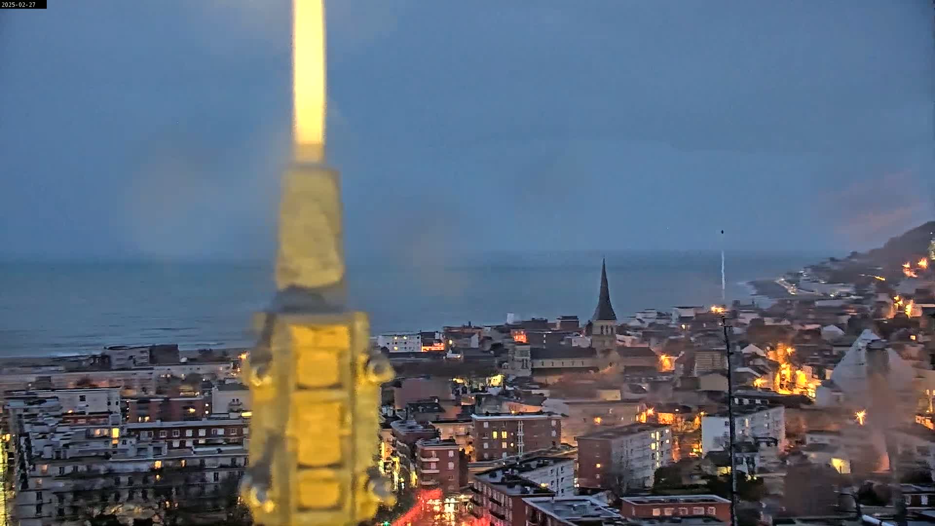 A coastal town at twilight is seen from a high vantage point, with a blurry, illuminated object in the foreground obscuring the view, under a mostly clear sky.