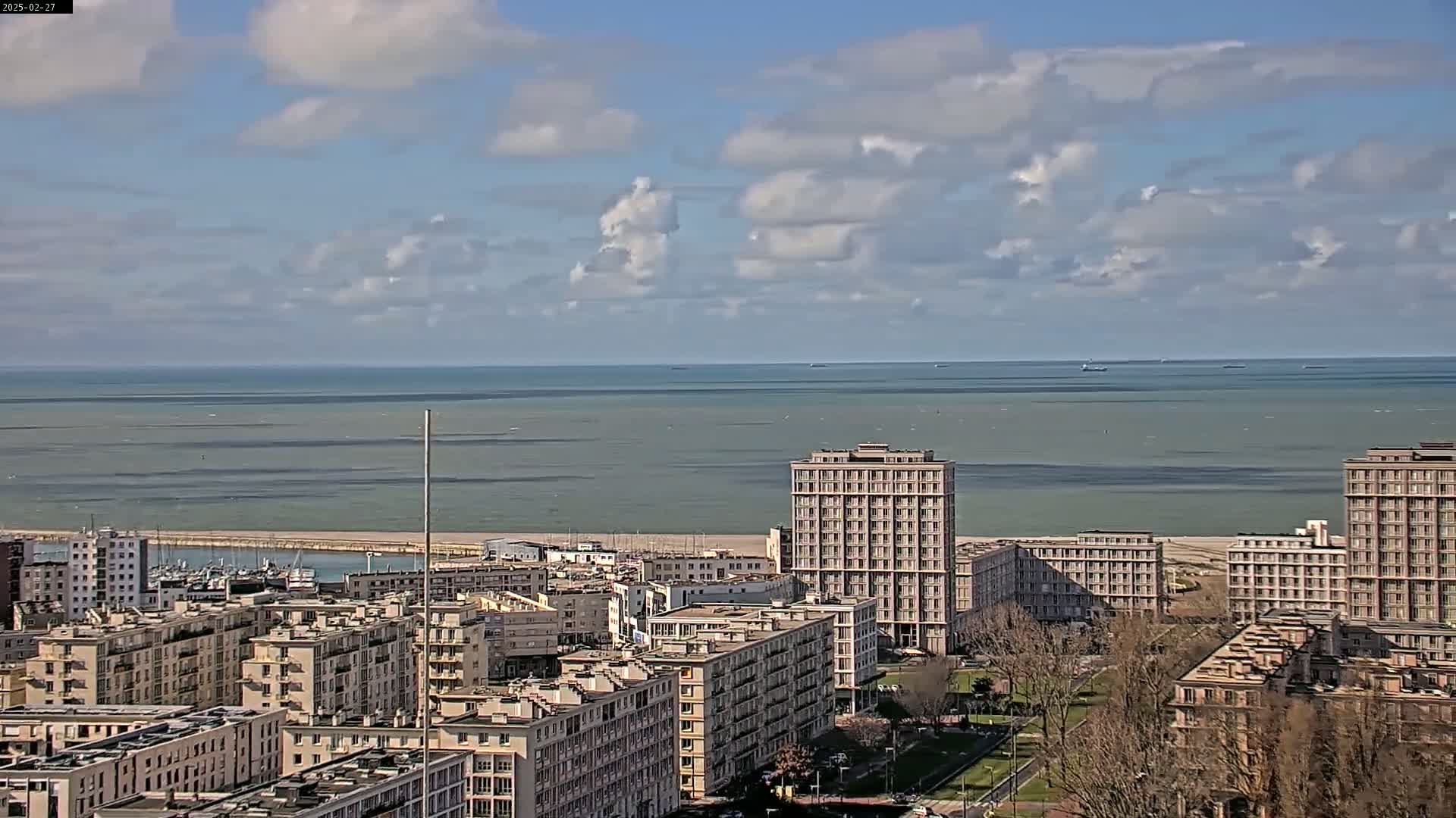 A coastal city with numerous apartment buildings is visible under a partly cloudy sky, overlooking a calm sea.