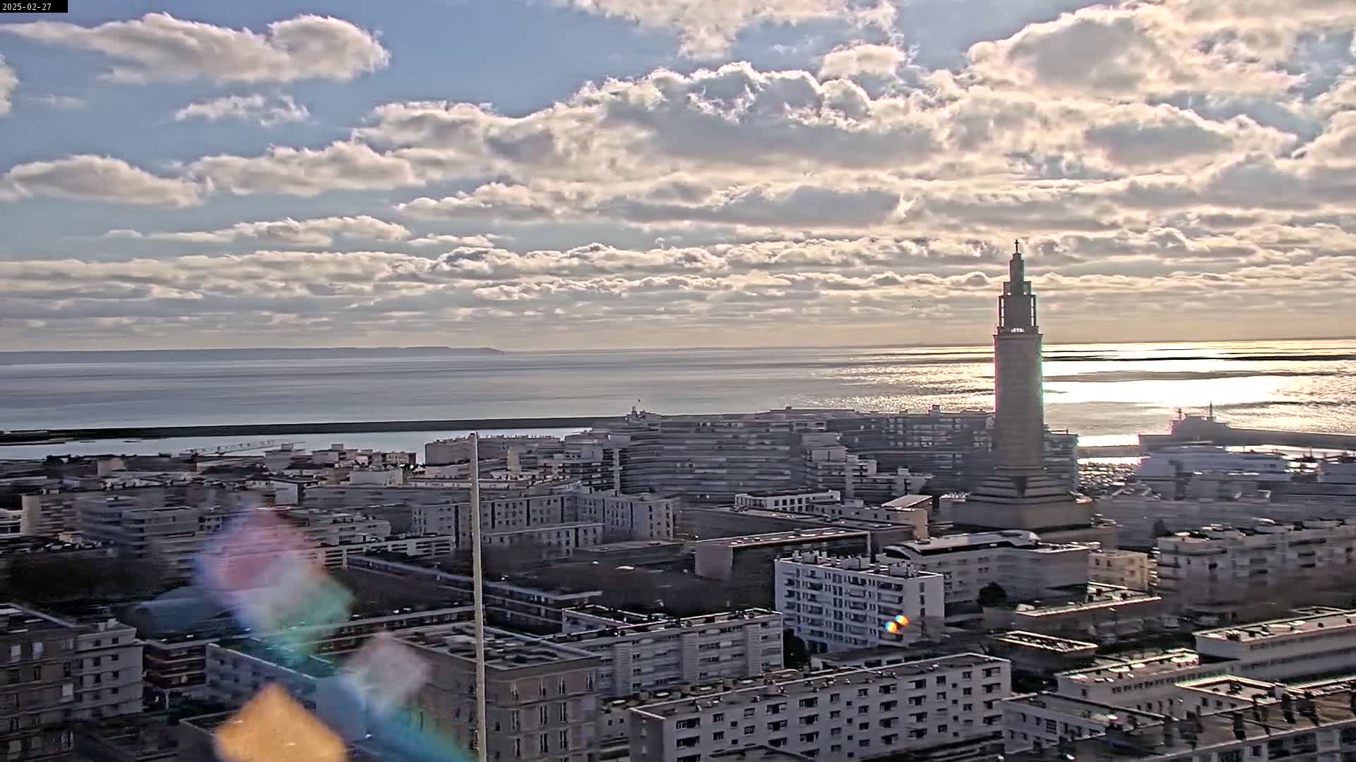 A coastal city with a tall tower is seen under a partly cloudy sky, with a calm body of water in the background.