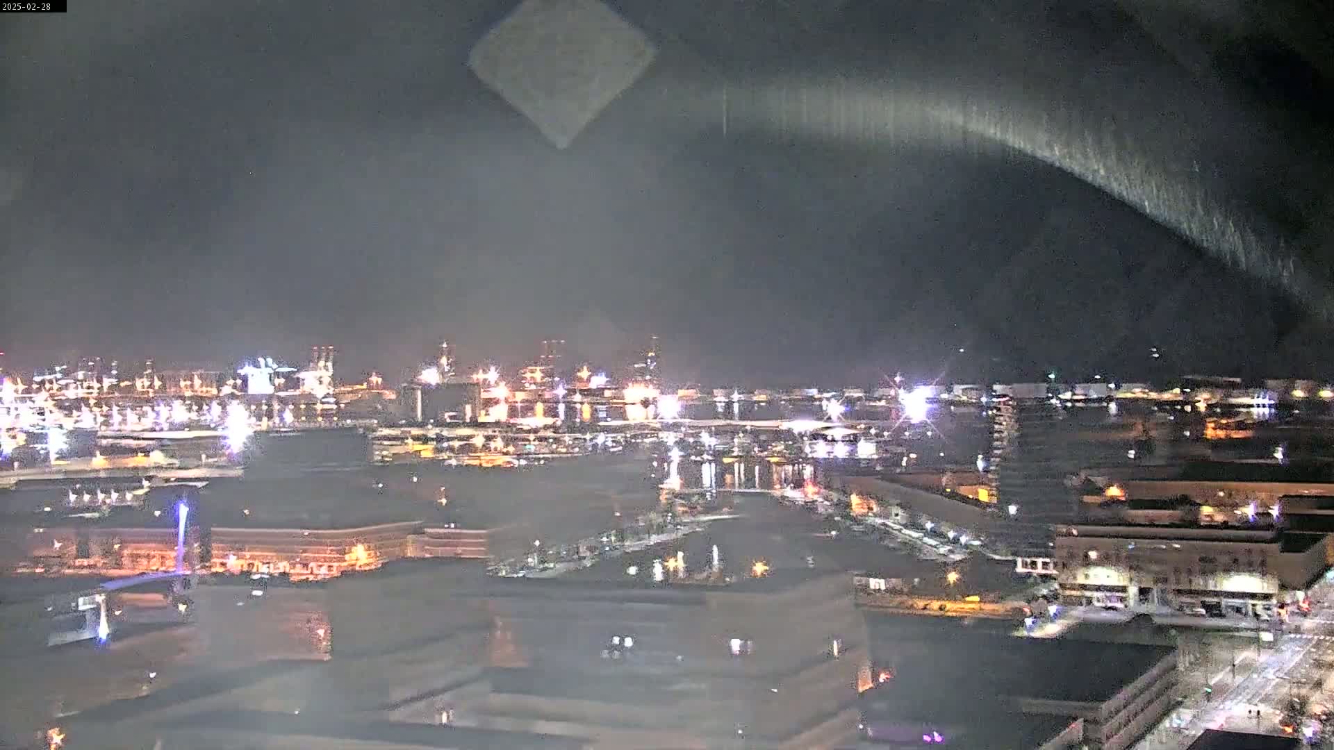 A nighttime view of a city's waterfront, featuring a harbor with many lights, and surrounding buildings, under a dark, mostly clear sky.