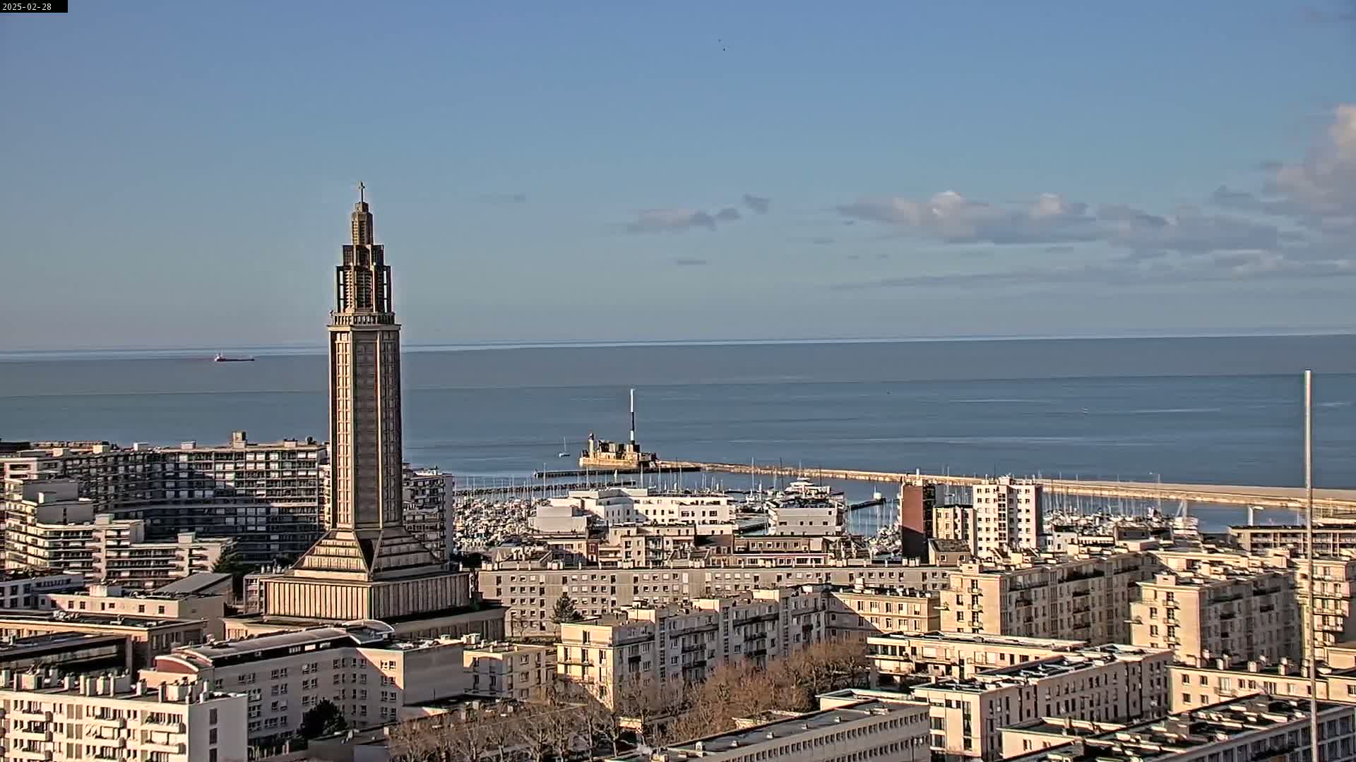 A tall, slender tower stands prominently in a cityscape overlooking a calm ocean under a mostly clear sky.