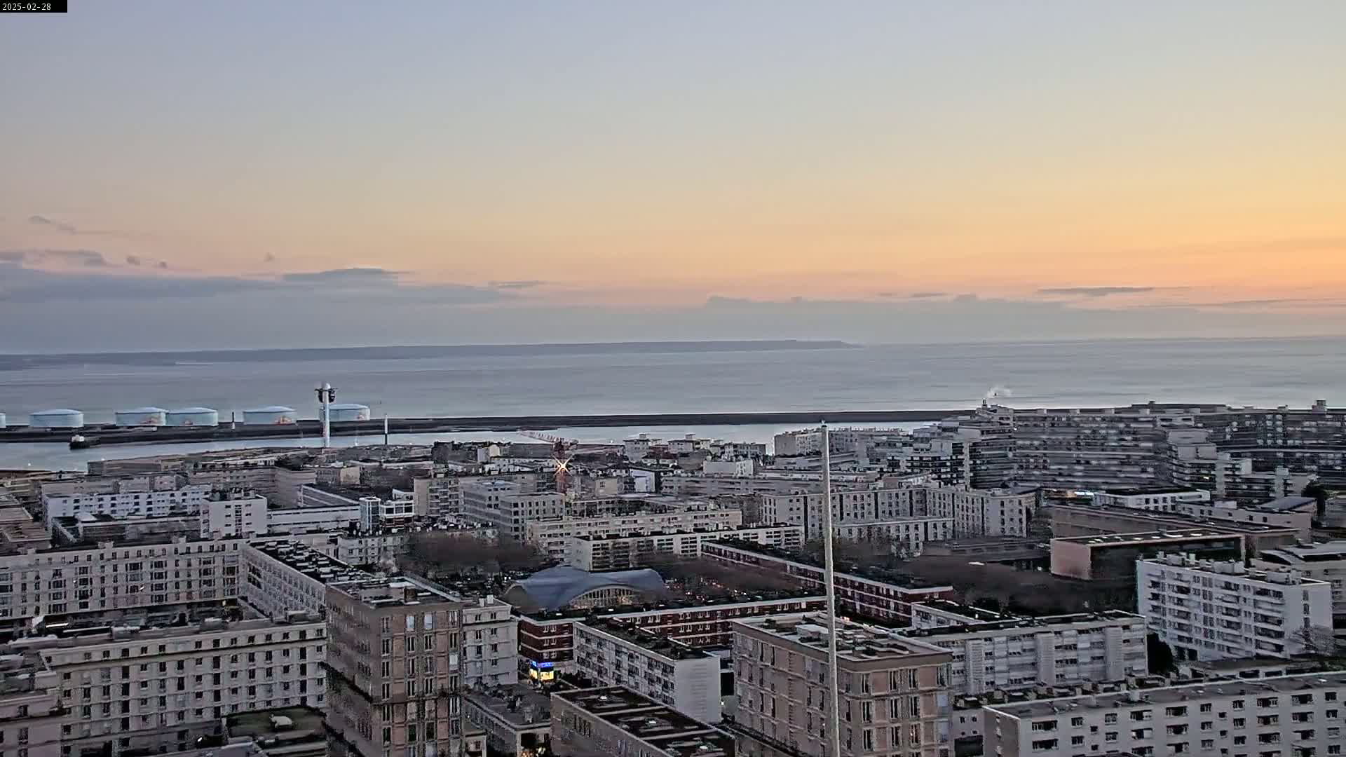 A sunset view of a city by the sea, showing a large expanse of water under a pale pink and orange sky.