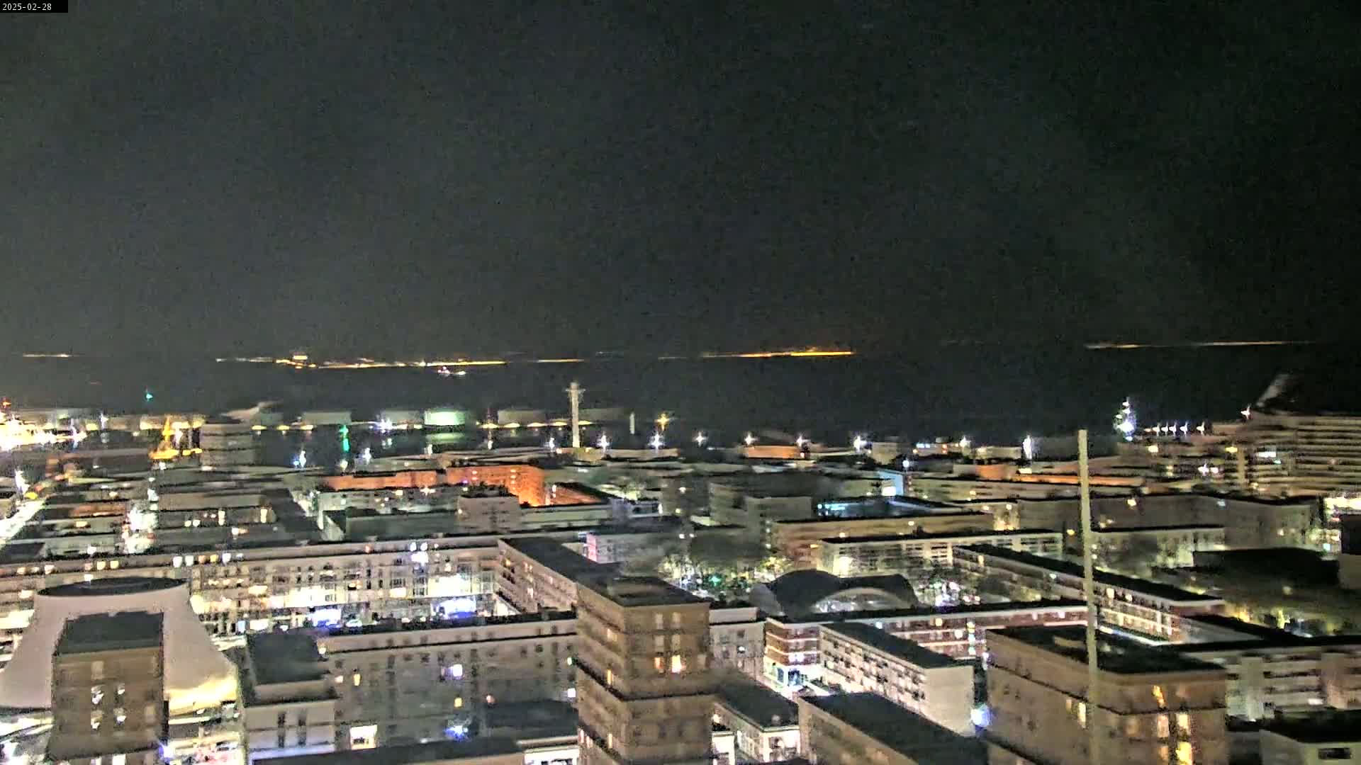 A nighttime aerial view of a city with a harbor and a body of water in the background under a clear, dark sky.