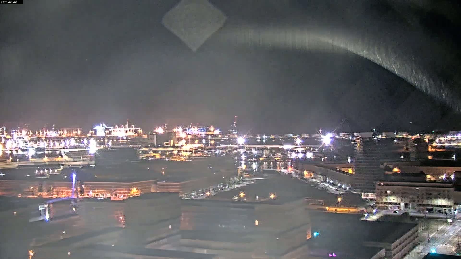 A nighttime, high-angle view of an industrial waterfront area with many lights, under a dark sky with a faint, arcing streak of light.