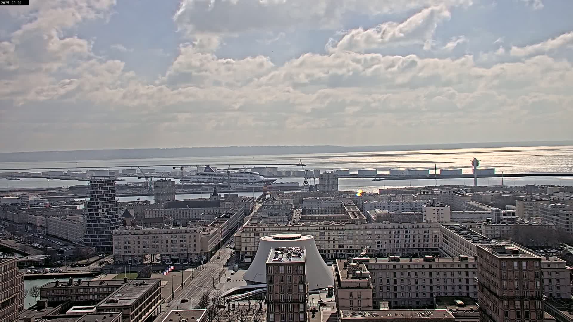 A partly cloudy day reveals a city harbor with several ships and docked tankers, stretching to a wide body of water under a mostly cloudy sky.