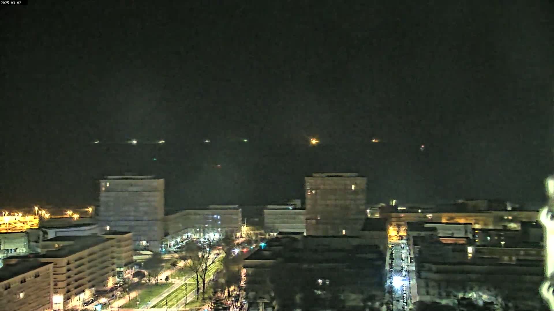 A nighttime view of a city skyline with several buildings illuminated, overlooking a dark body of water with several distant lights visible on the horizon under a clear, dark sky.