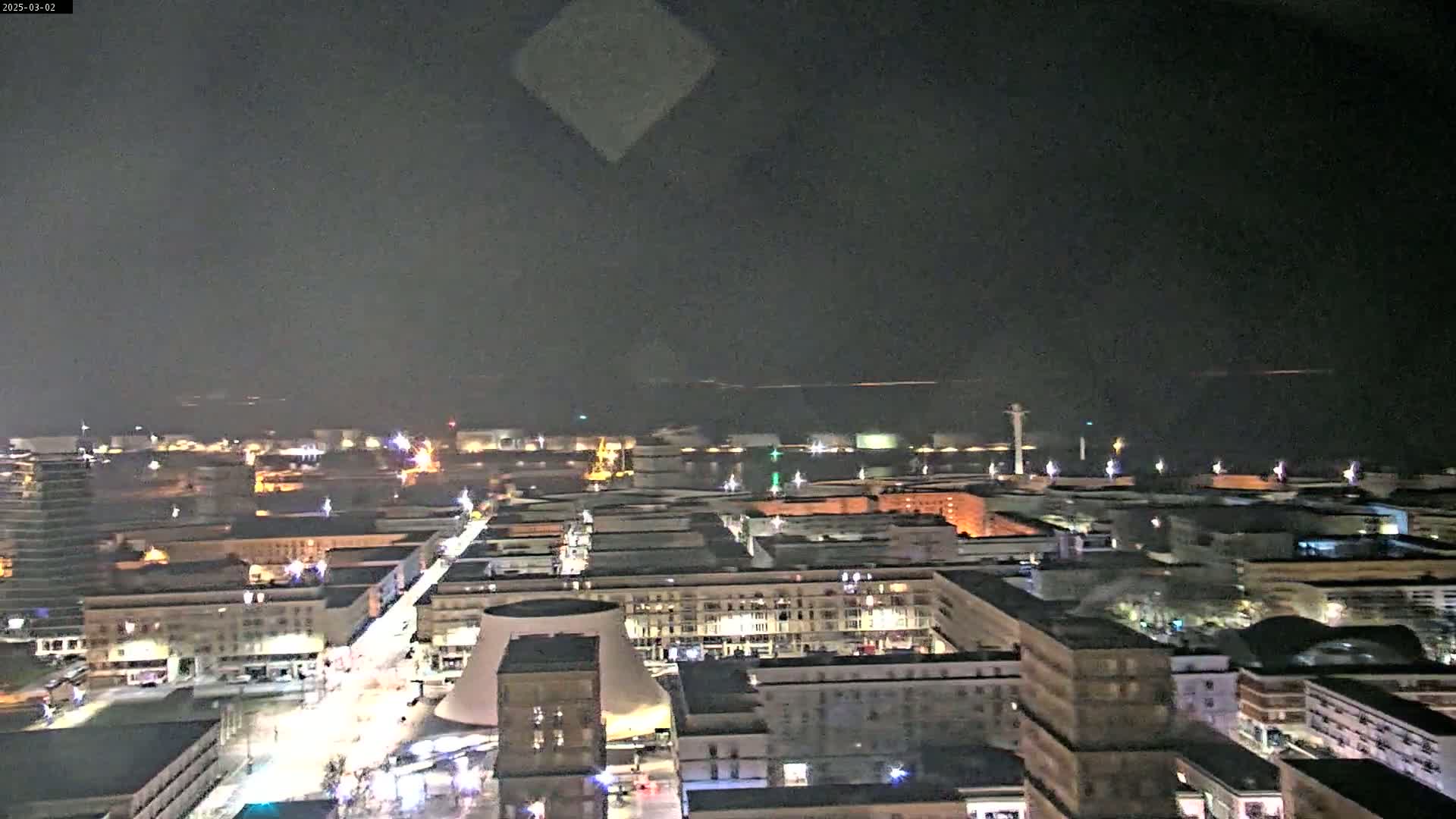 A nighttime aerial view of a city with illuminated buildings and streets, extending to a dark, calm body of water in the distance.