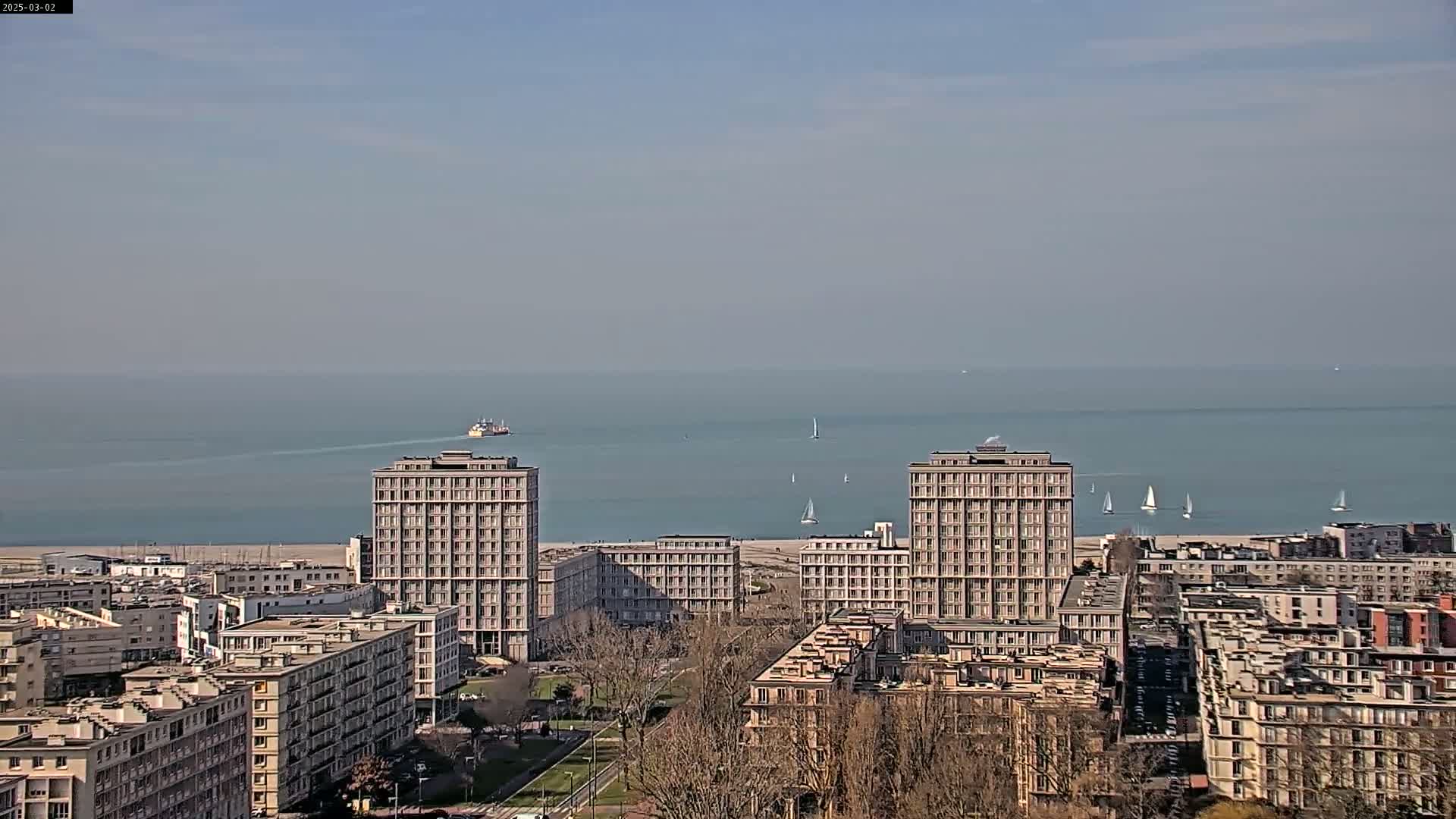 A hazy, sunny day reveals a cityscape of several multi-story buildings next to a calm sea with a few sailboats and a distant ship.