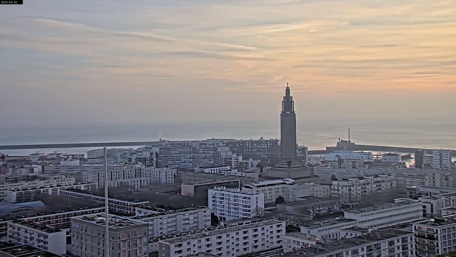 A coastal city with numerous apartment buildings and a tall tower is seen at sunrise under a hazy, pale sky.