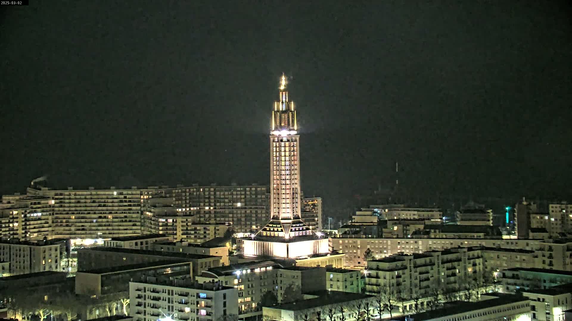 A tall, illuminated tower stands prominently in a city at night, surrounded by residential buildings under a dark sky.