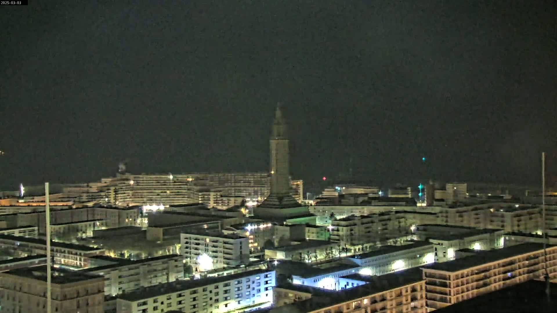 A nighttime aerial view of a city with a tall tower in the center, under a dark, cloudy sky.