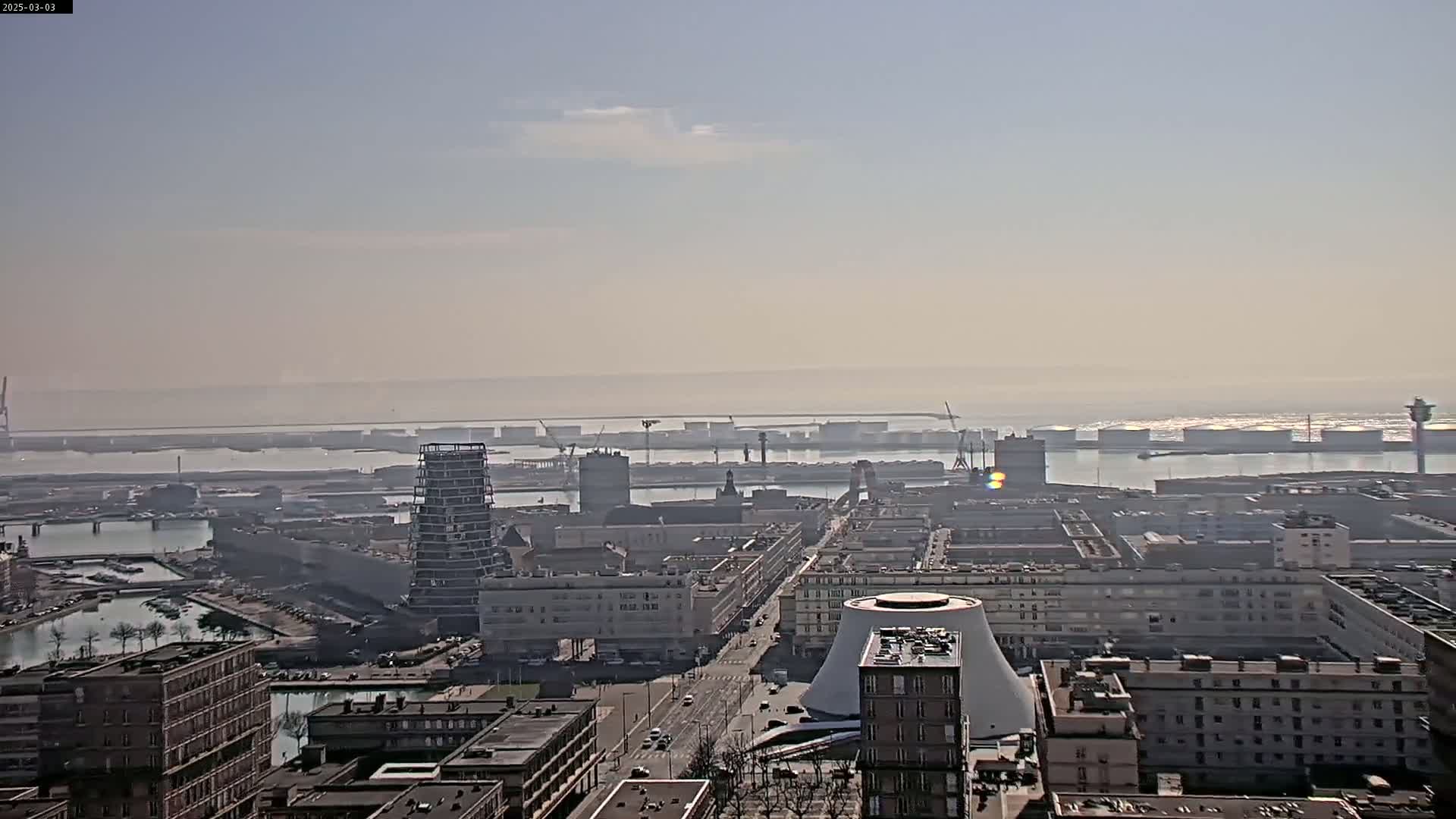 A hazy, sunny day reveals a sprawling cityscape overlooking a harbor filled with ships and industrial structures.
