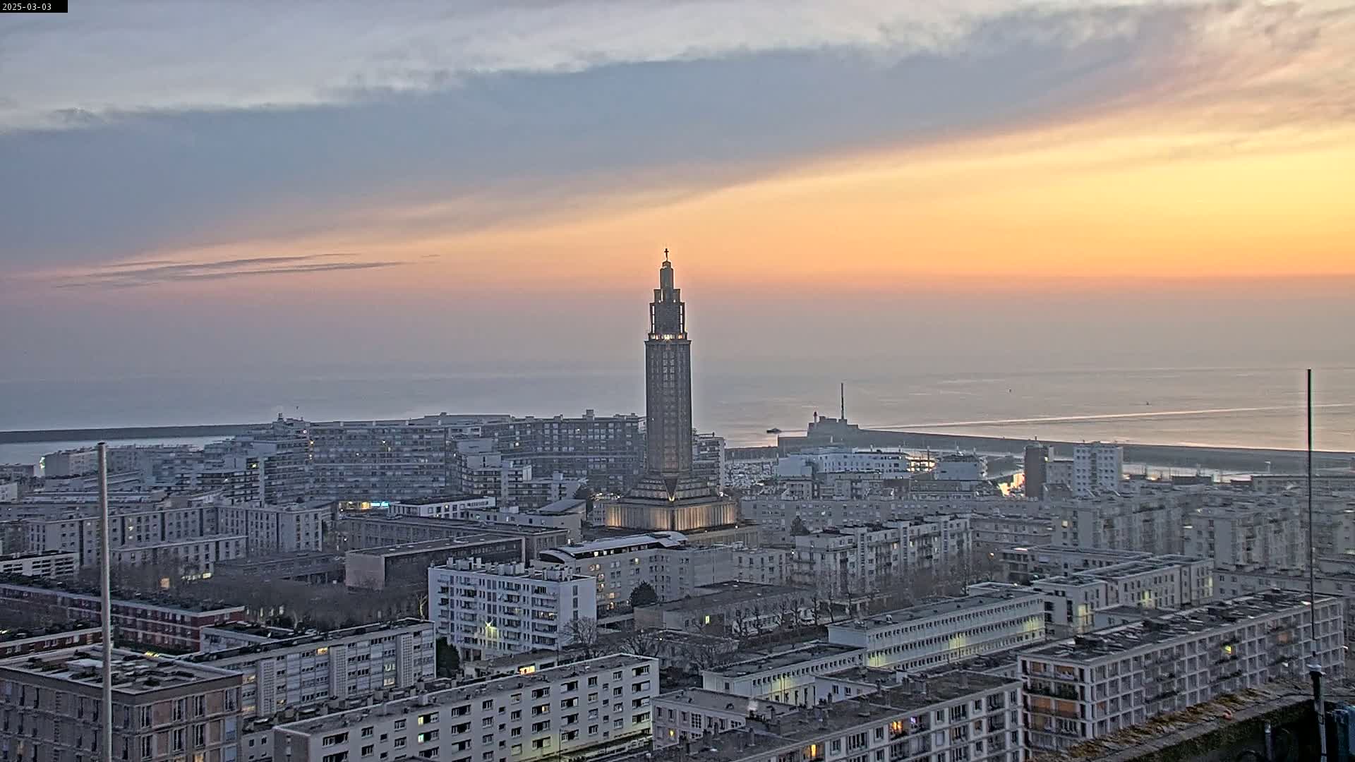 A coastal city at sunset, featuring a tall, slender tower at its center, under a mostly clear sky with a band of pale orange and yellow hues near the horizon.