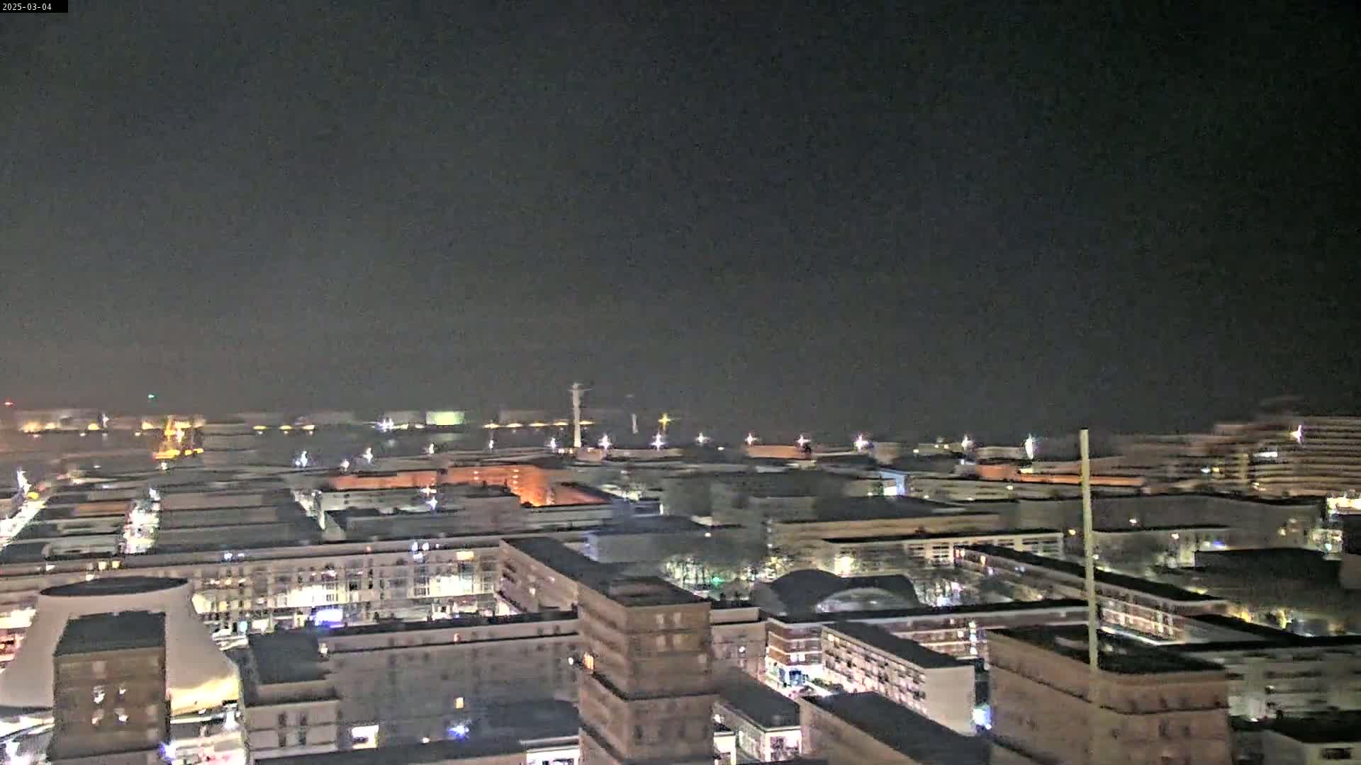 A nighttime, high-angle view shows a city's waterfront area with many buildings illuminated, and a body of water in the distance.