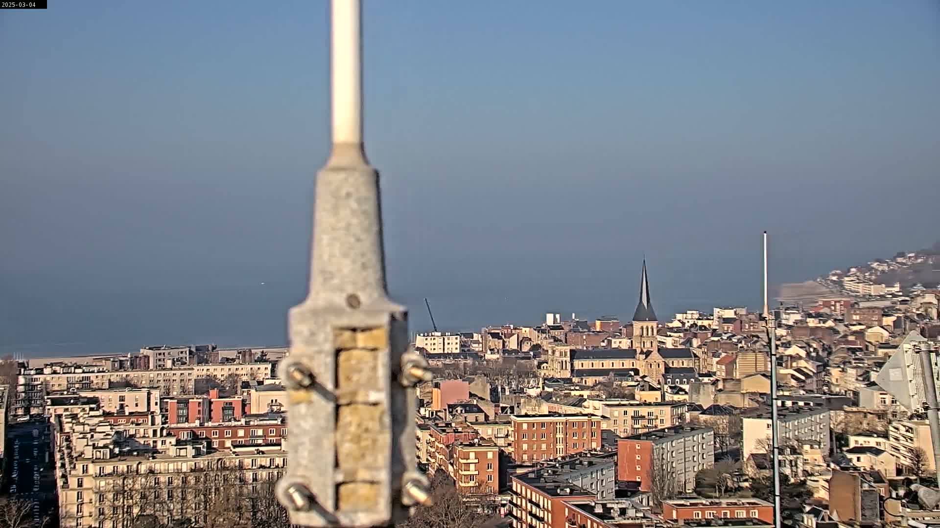 A hazy, sunny day reveals a coastal city with a church steeple visible among many buildings, all viewed from a high vantage point behind a metal pole.