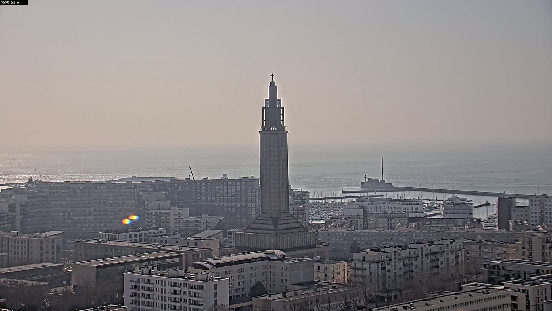A tall, slender tower stands in the center of a city overlooking a calm sea on a hazy day.