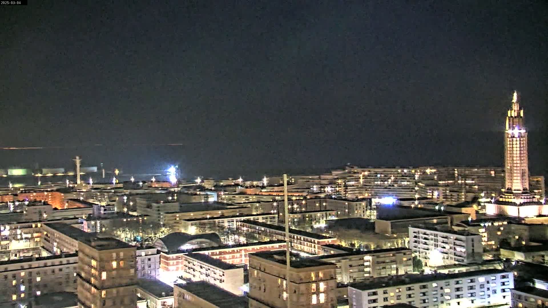 A nighttime aerial view of a city with a tall tower illuminated, overlooking a dark body of water under a clear sky.