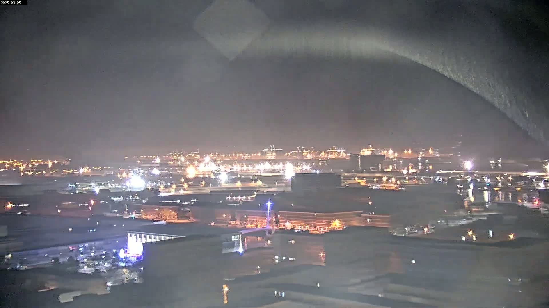 A nighttime, possibly rainy, view of a city's waterfront and industrial area, illuminated by numerous lights, with a hazy or foggy atmosphere.