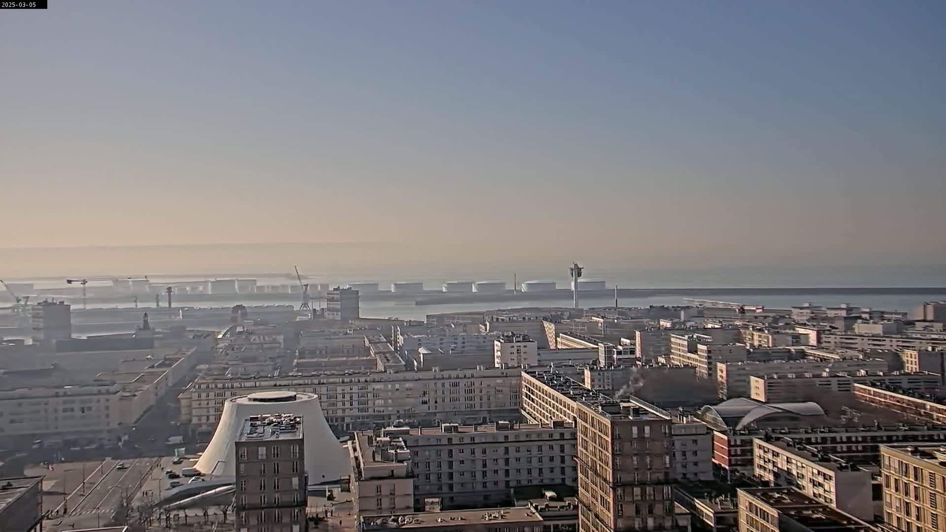 A hazy, sunny day reveals a cityscape overlooking a harbor filled with large vessels and industrial structures.