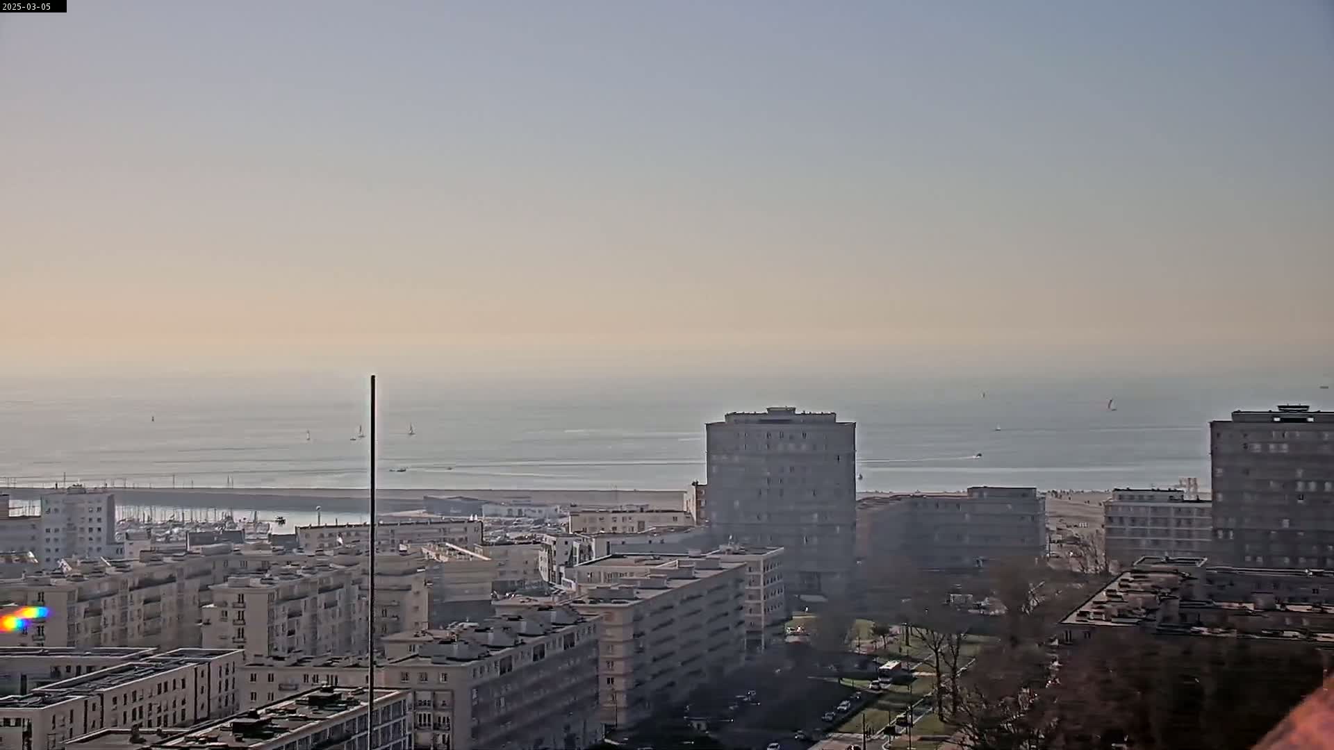 A hazy, calm ocean is visible in the distance behind a cityscape of multi-story buildings.