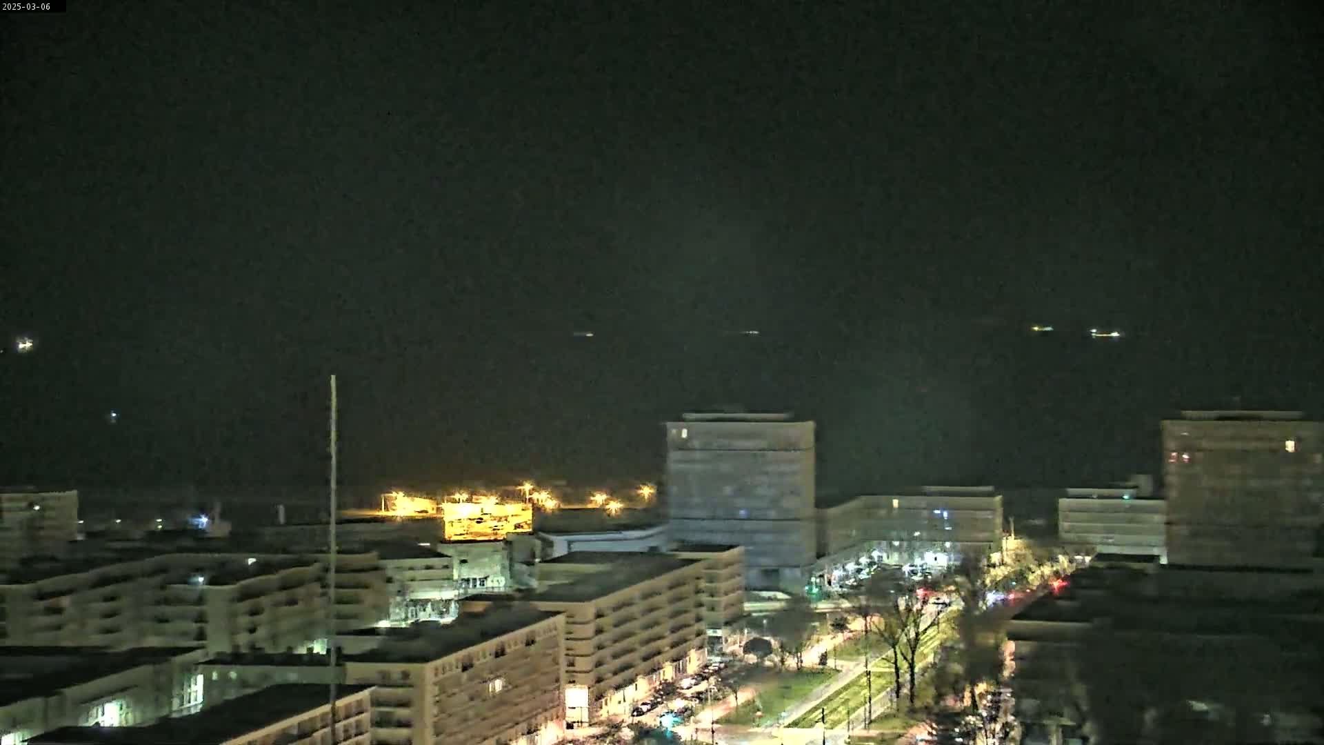 A nighttime aerial view of a city waterfront shows several multi-story buildings with illuminated windows, a street with moving vehicles, and distant lights on the water.