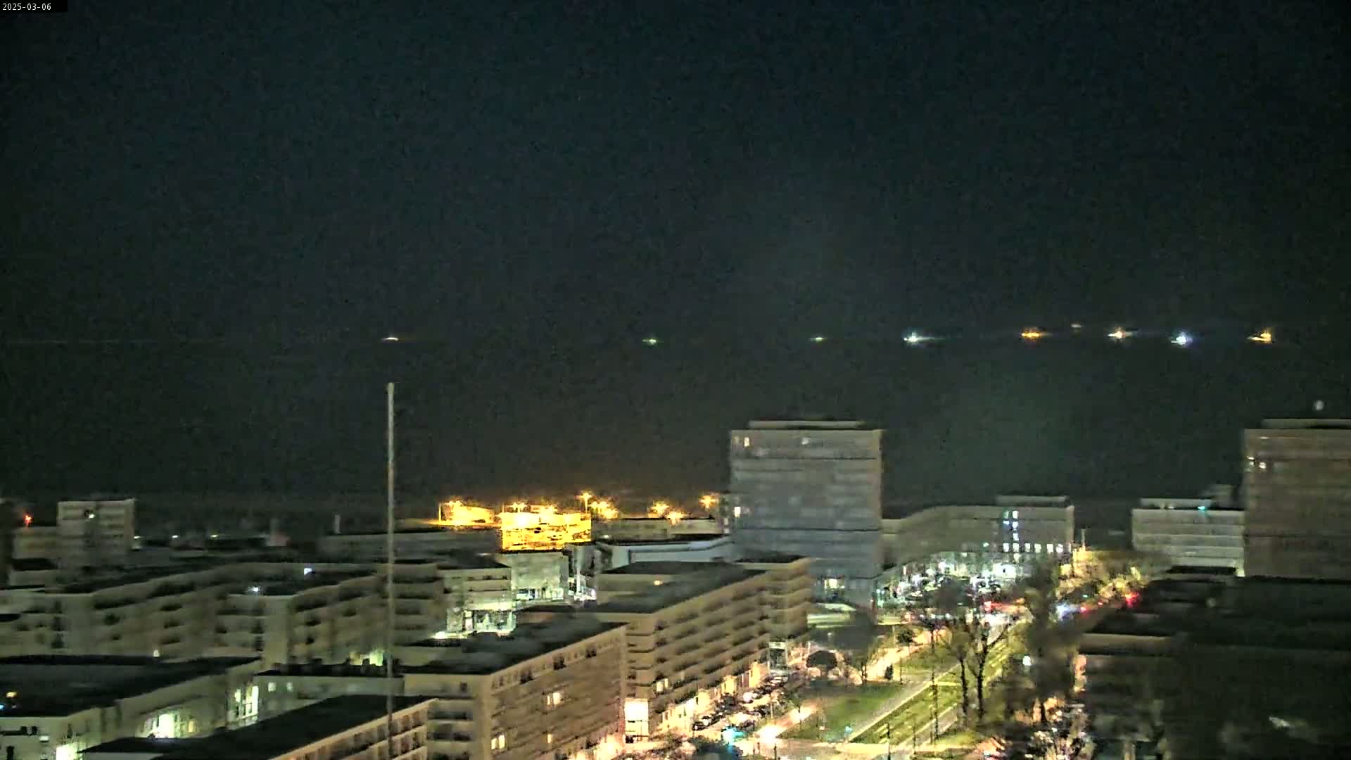 A nighttime view of a city waterfront shows several lights on the water and illuminated buildings along a street.