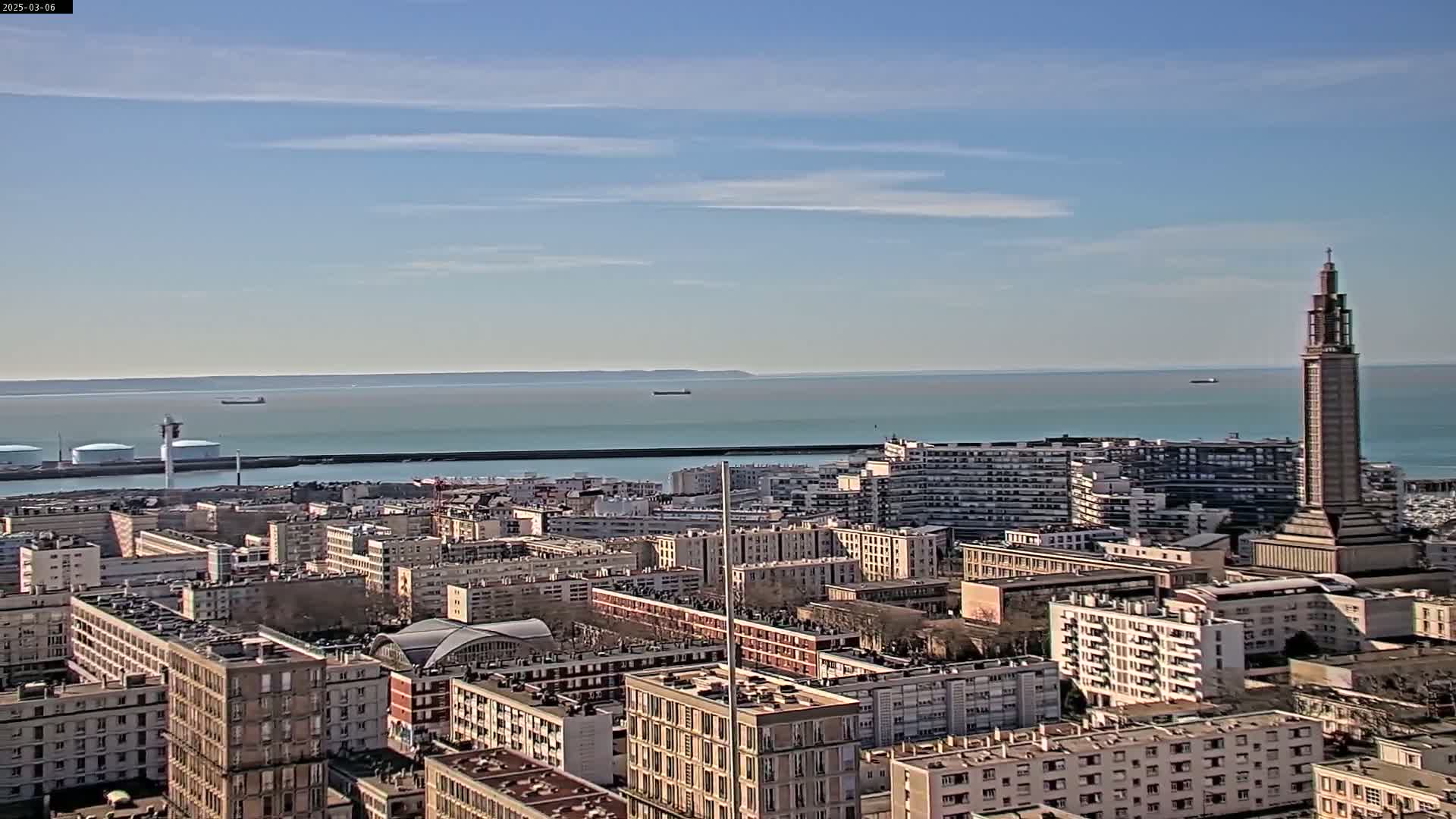 A coastal city with numerous buildings and a tall tower is visible under a mostly clear, sunny sky, with calm water and a few ships in the distance.