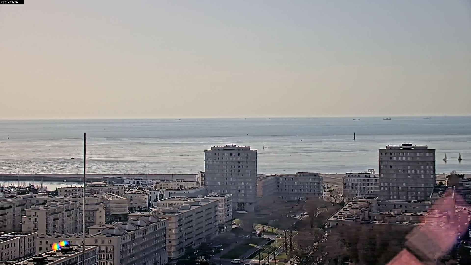 A calm, light-grey sea stretches to the horizon beyond a coastal city of mostly multi-story apartment buildings.