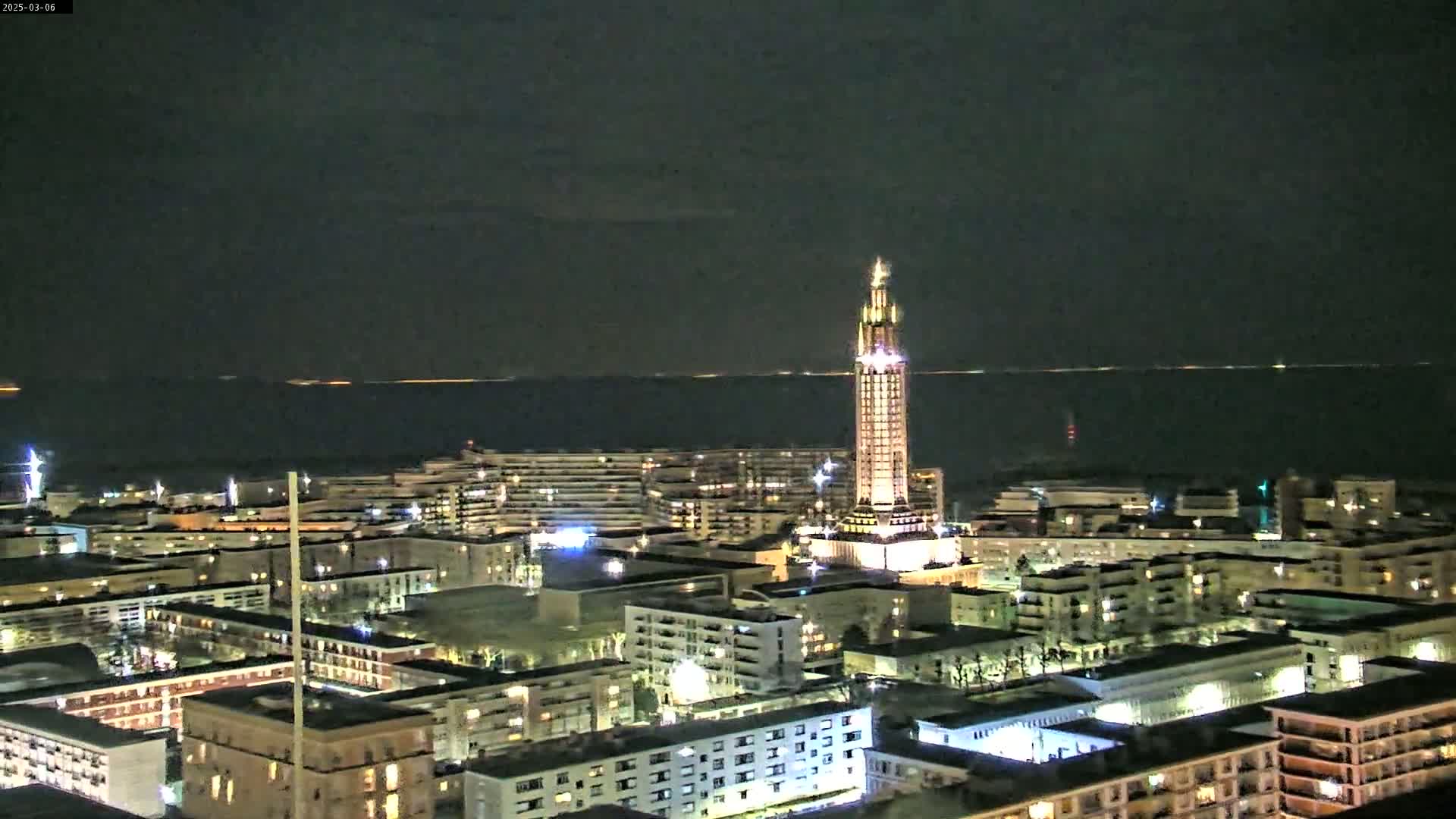 A nighttime aerial view of a city illuminated with lights, featuring a tall, slender tower, overlooking a dark body of water under a clear night sky.