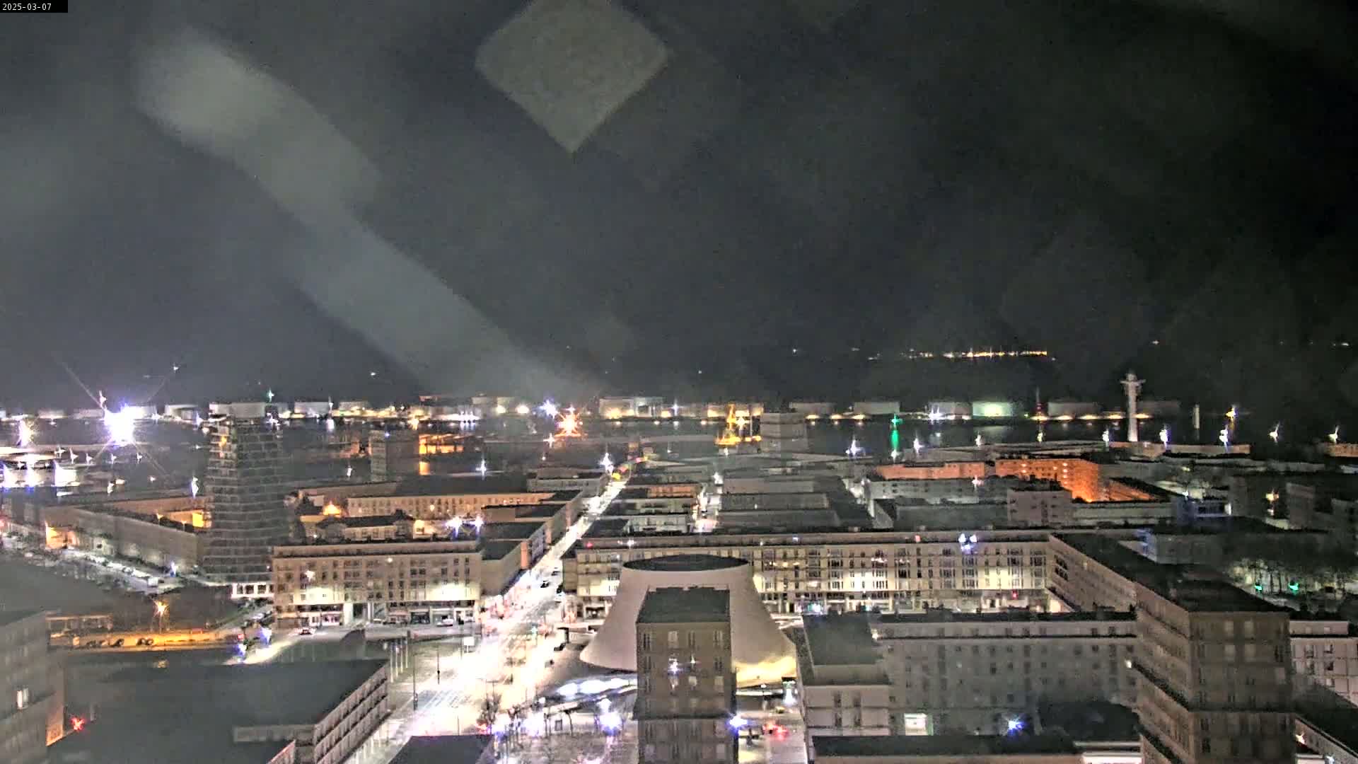 A nighttime aerial view of a city illuminated by streetlights, showing a waterfront area and a distinctive domed building, under a dark, slightly hazy sky.