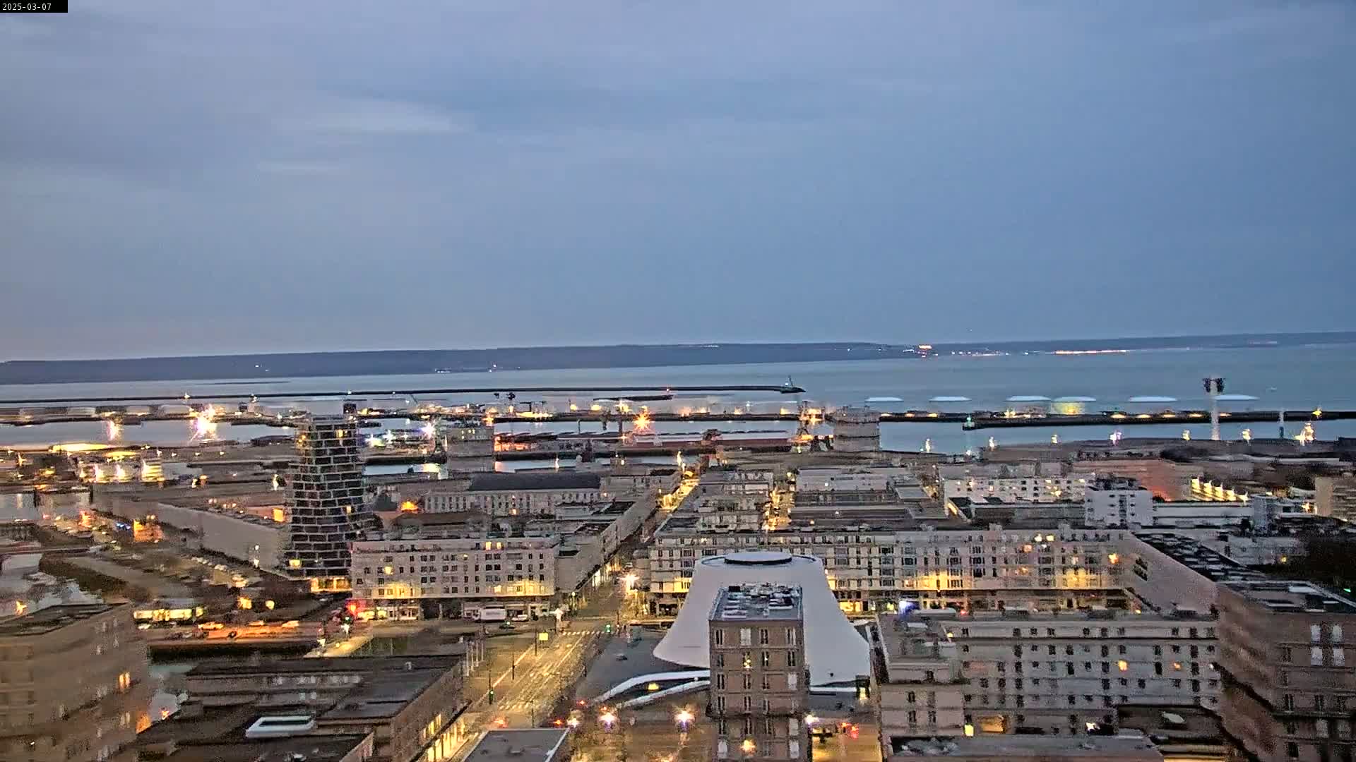 A nighttime, high-angle view of a city's harbor and residential area under a dusky sky, with city lights illuminating the buildings and the port.