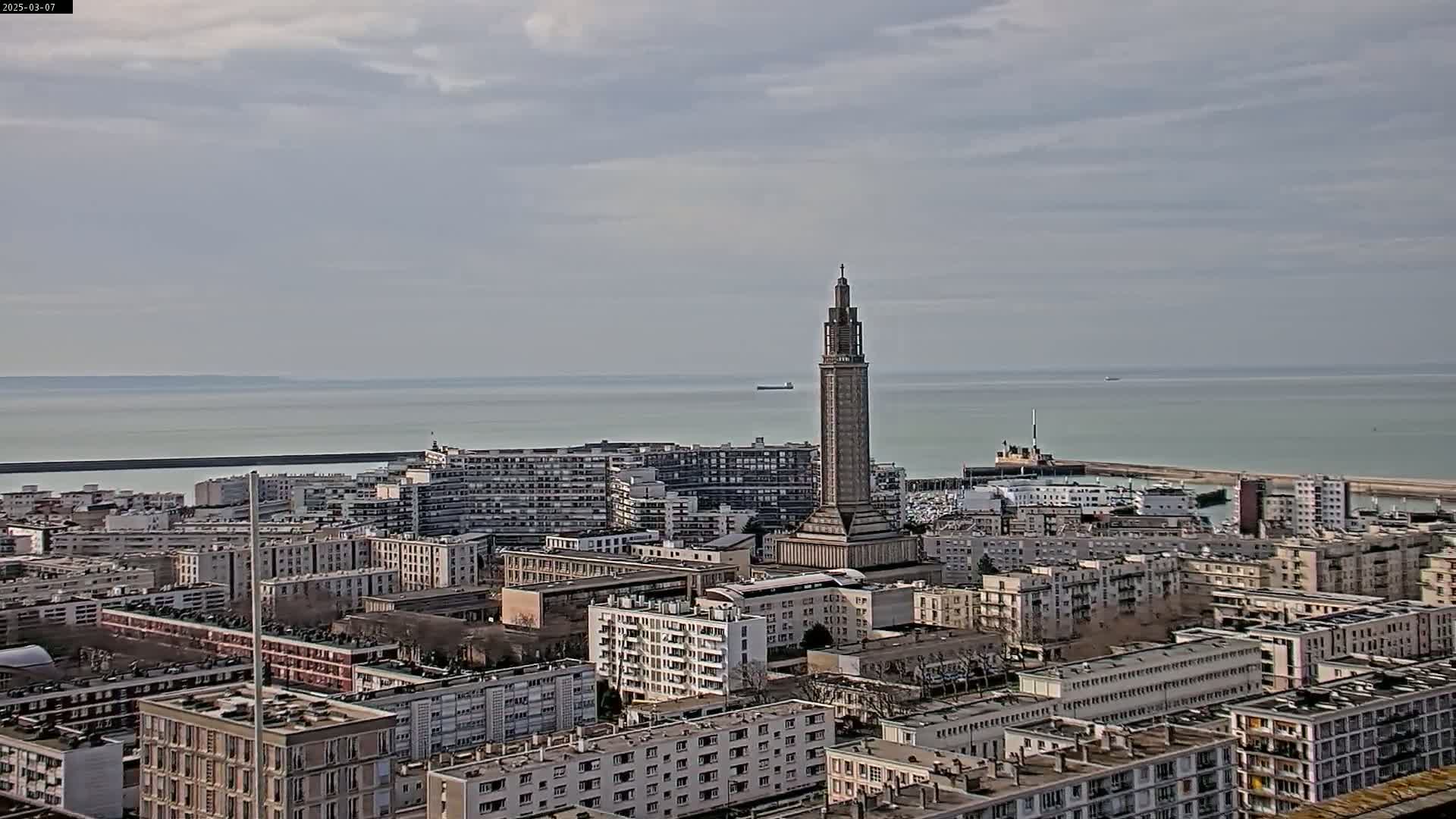 A coastal city with numerous apartment buildings, a tall tower, and a harbor is visible under an overcast sky.