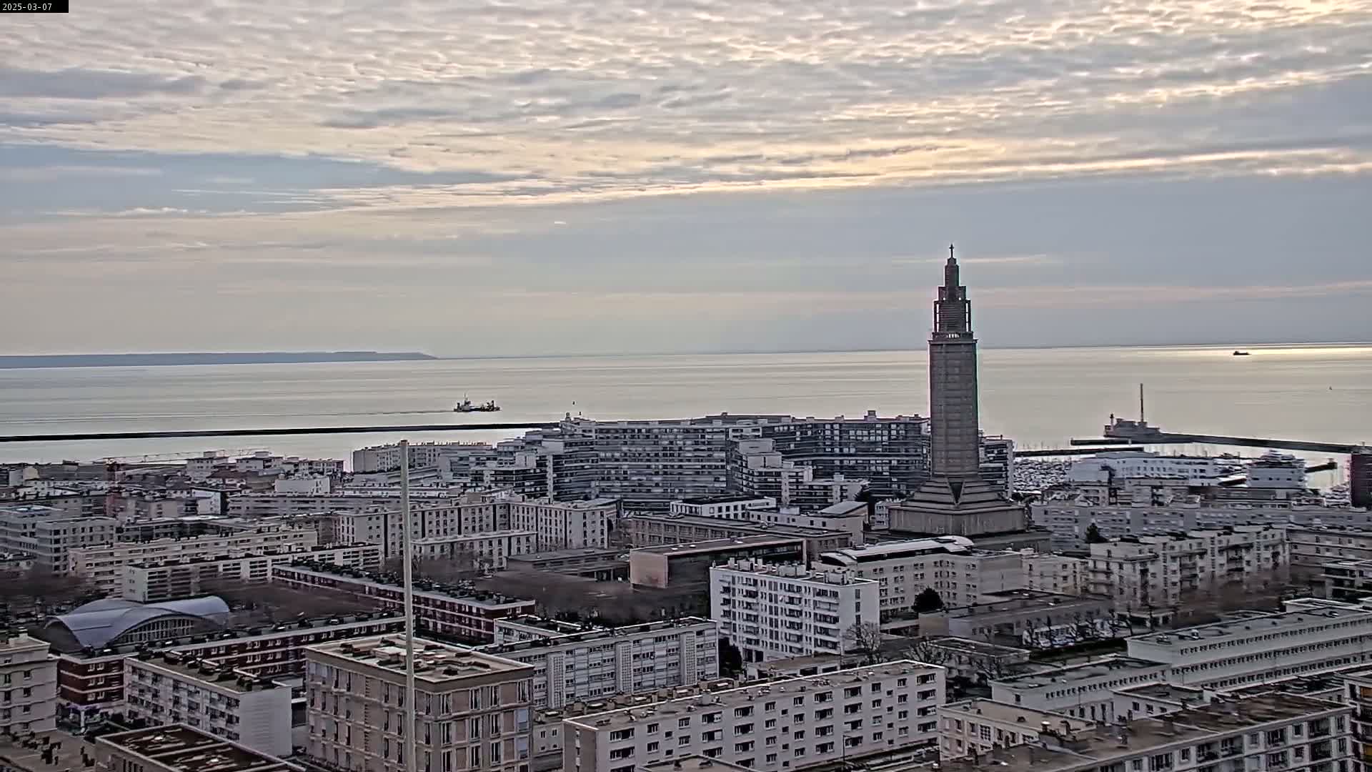 A coastal city with a tall tower is visible under a cloudy sky, with calm water and a distant shoreline in the background.
