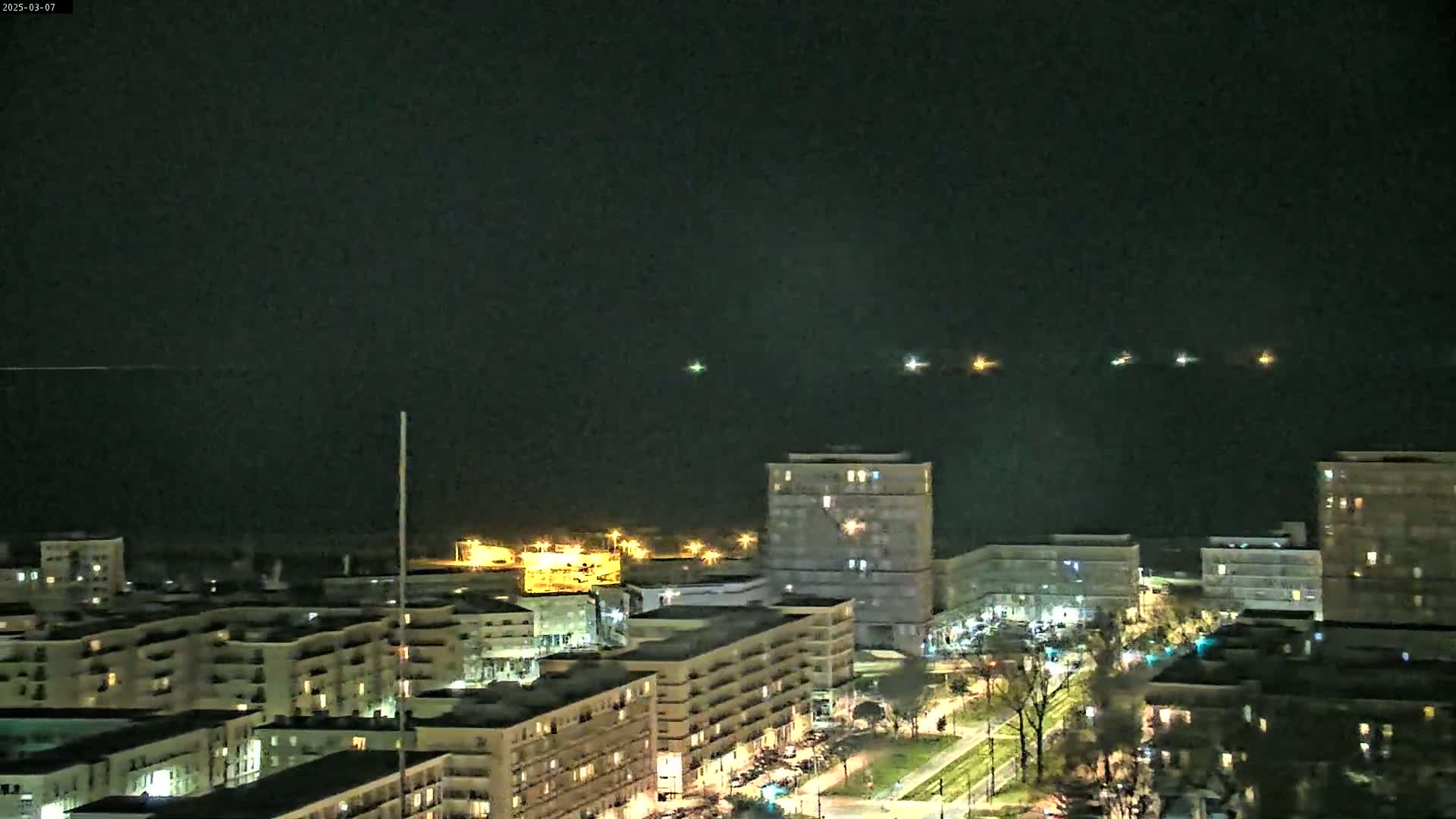 A nighttime view of a city with several multi-story buildings illuminated,  a body of water visible in the distance with faint lights, under a dark, clear sky.