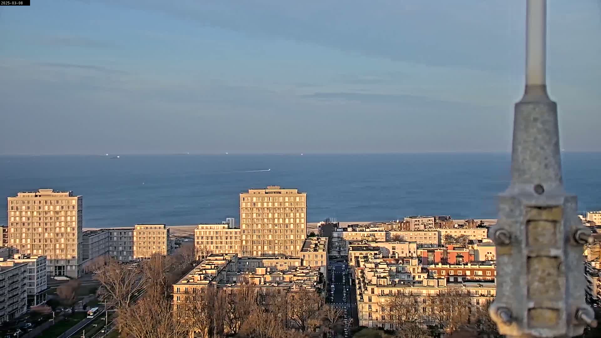 A calm ocean is visible in the background, beyond a city of low-rise buildings and several high-rise apartment buildings under a clear, light-blue sky.