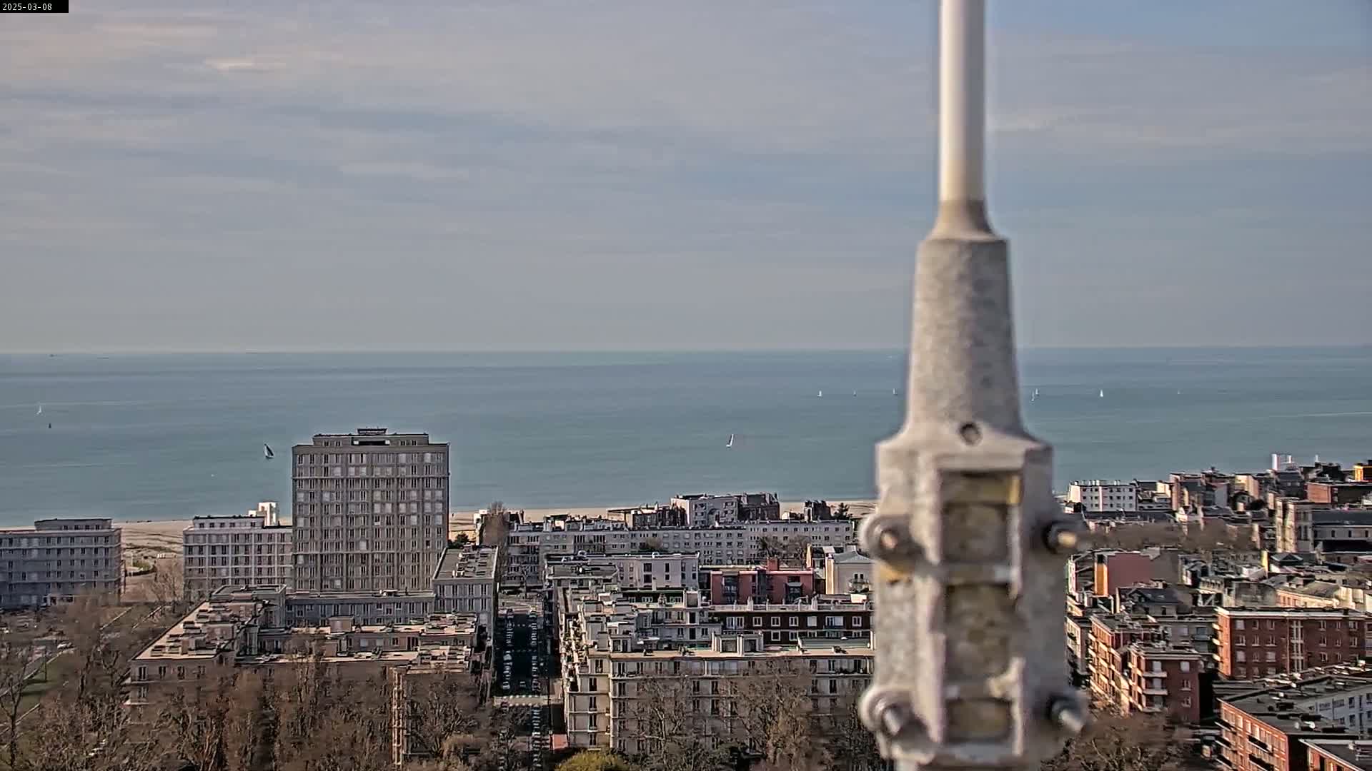 A coastal city viewed from above on a partly cloudy day shows a calm sea with sailboats in the distance, buildings, and a tall structure partially obscuring the view.