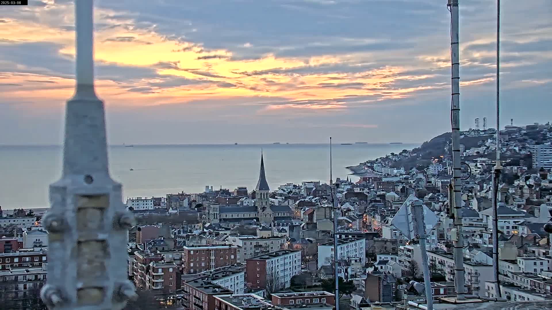 A coastal city with a church spire is seen at sunset under a partly cloudy sky, as viewed from a high vantage point.