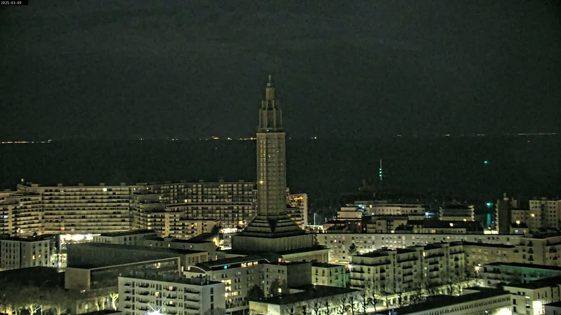 A tall, slender tower stands in the center of a city at night, overlooking a dark body of water under a mostly clear sky.