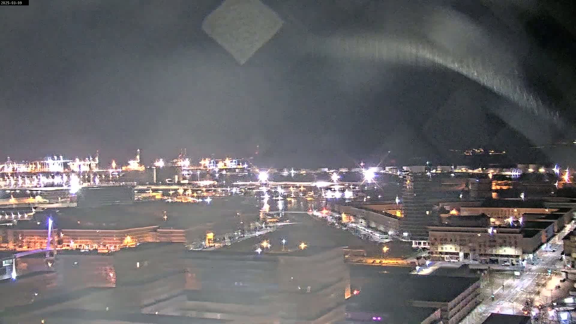 A nighttime view of a city's waterfront and industrial area, with lights illuminating buildings and ships under a dark, possibly cloudy sky.