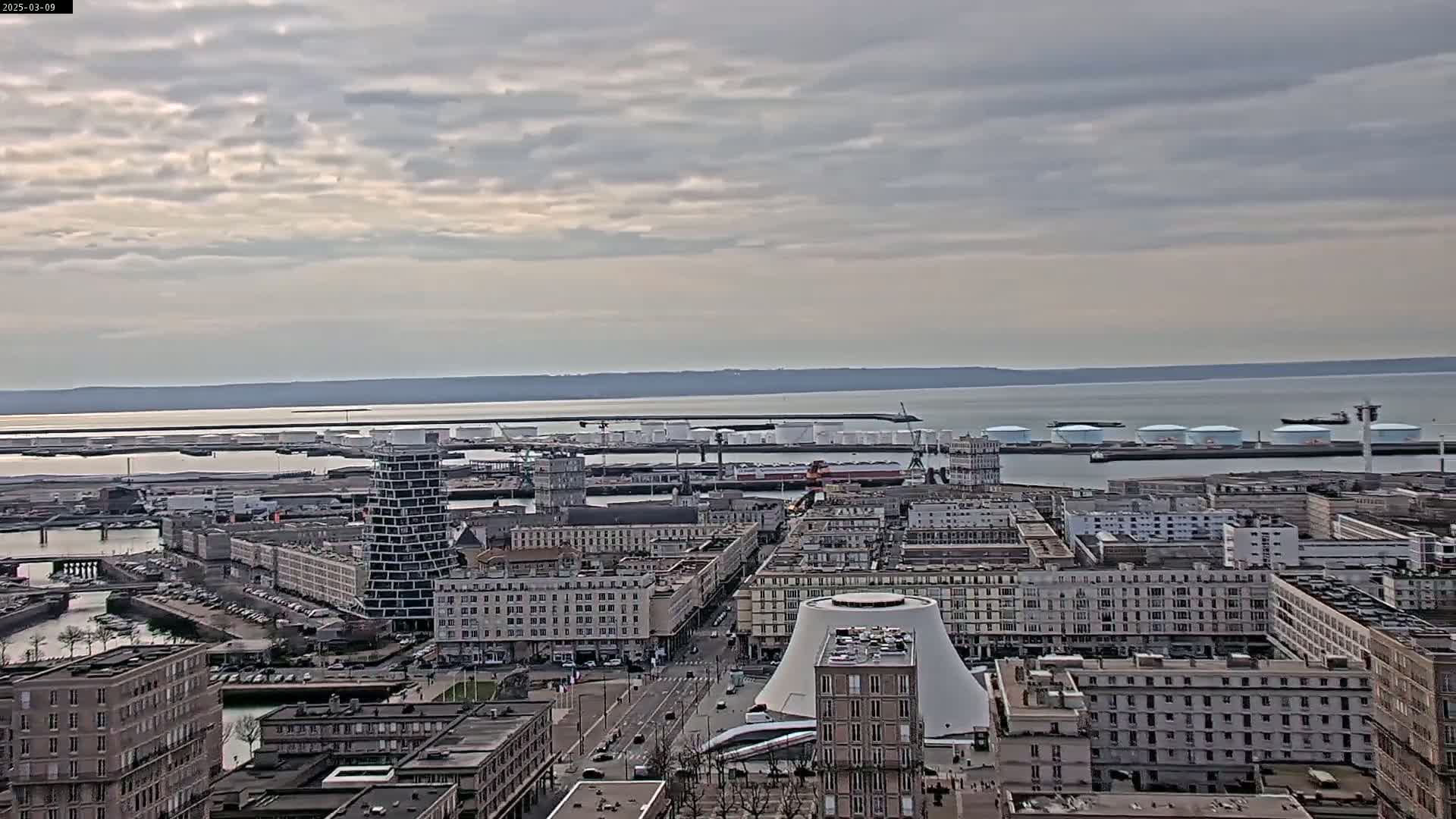 A cityscape with a harbor, featuring numerous buildings, a tall slender tower, and a large dome-shaped structure, all under a cloudy sky.