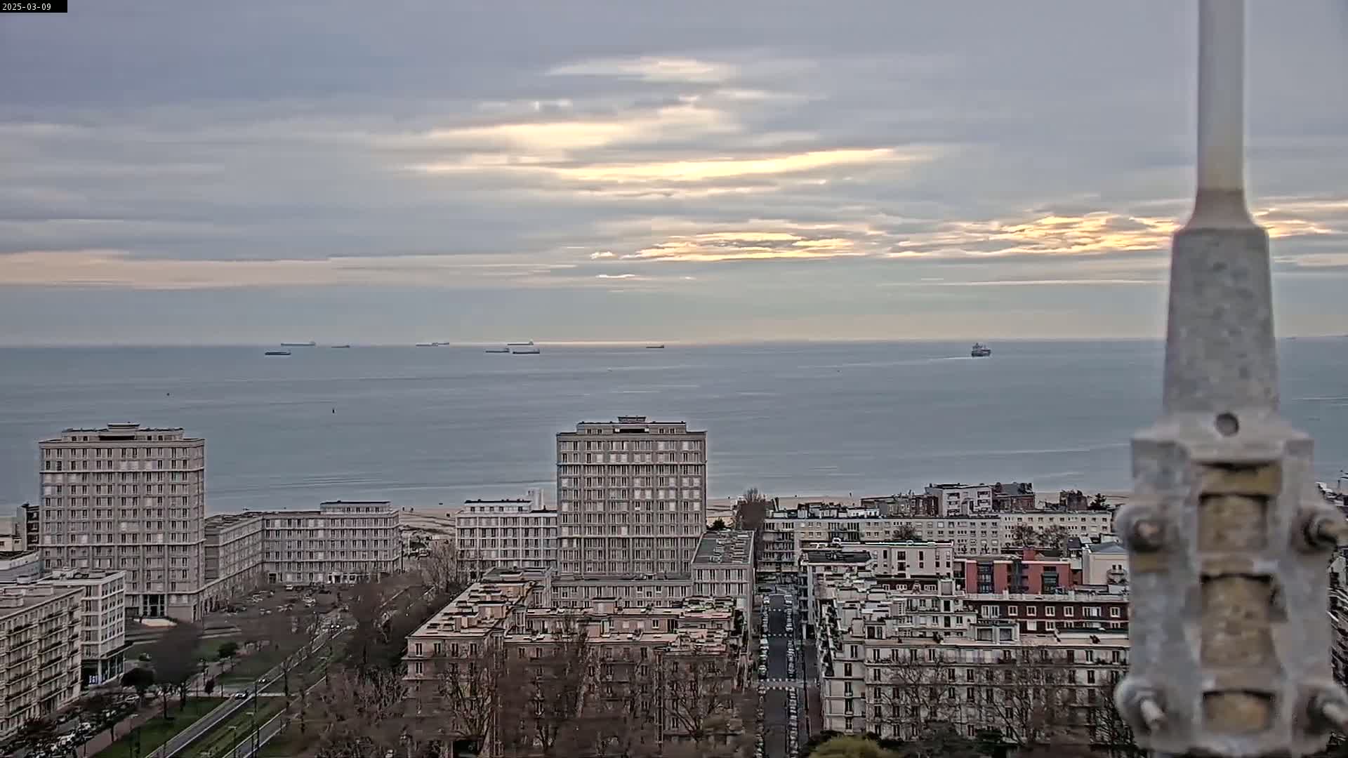 A cityscape of multi-story buildings overlooks a calm ocean with several ships in the distance under a partly cloudy sky.