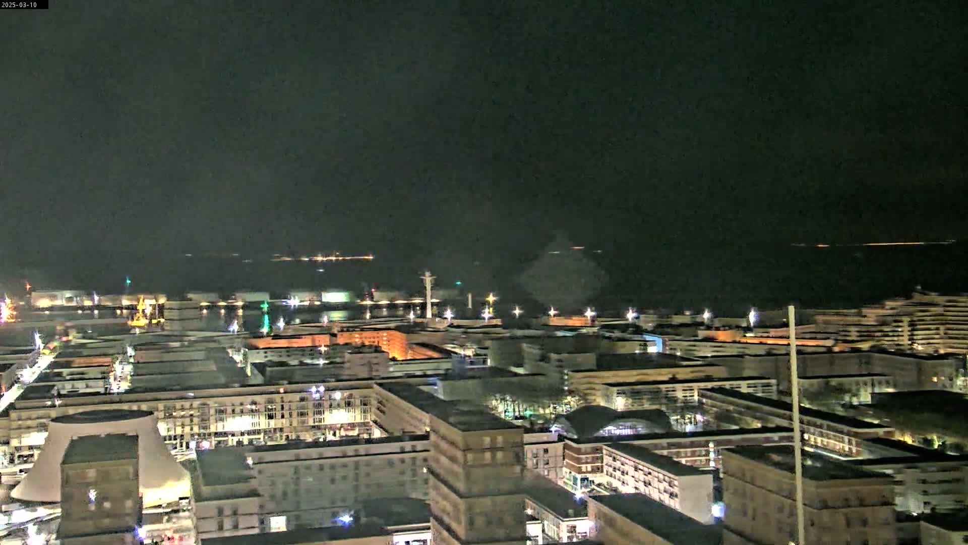 A nighttime aerial view of a city harbor with many buildings illuminated, overlooking a dark body of water under a clear sky.