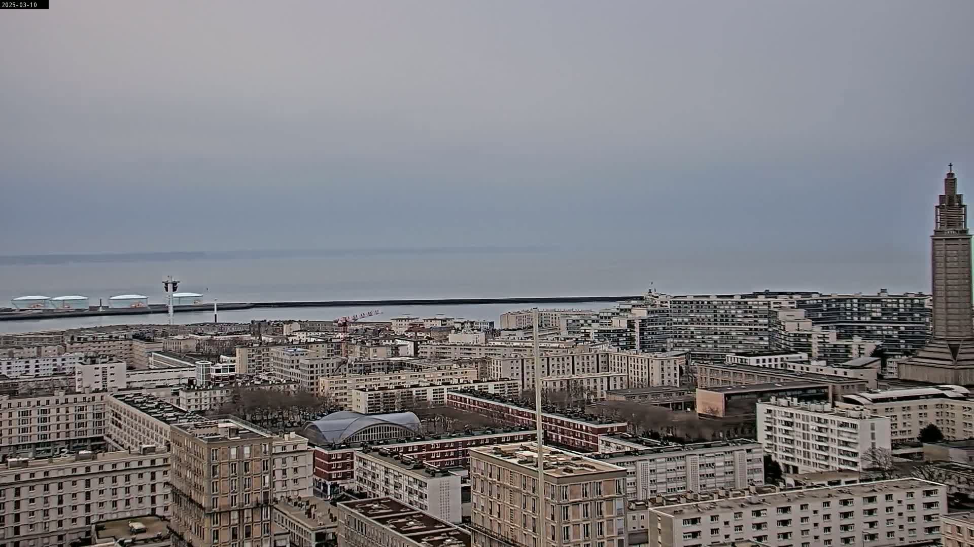An overcast day reveals a cityscape extending to a calm body of water, featuring a variety of buildings including high-rises and a prominent tower.